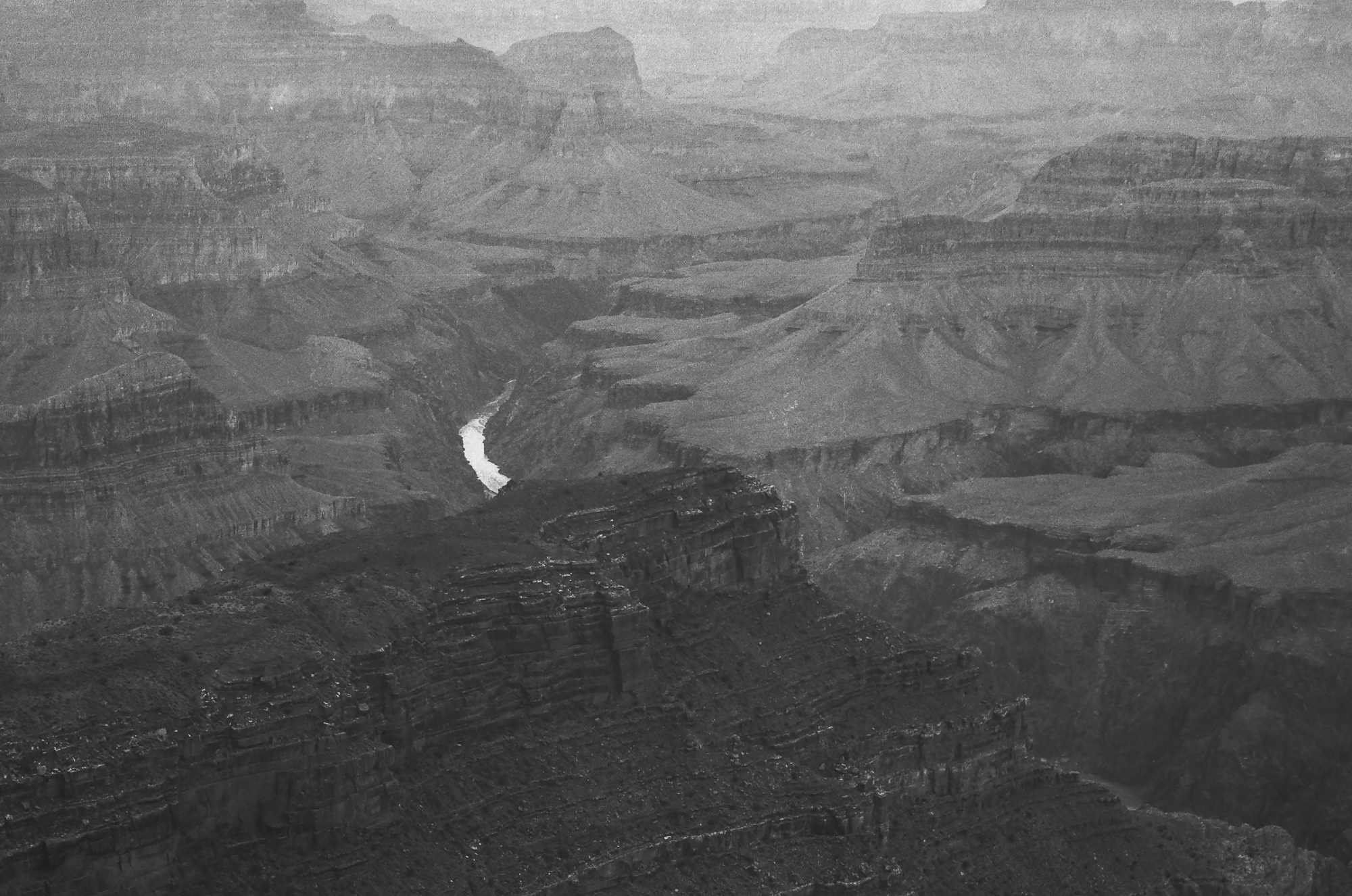 A high, hazy black and white view into the Grand Canyon, with receding ridges and terraced cliffs stacked from dark foreground rock to pale distant mesas. A narrow bend of the Colorado River reads as a bright white ribbon at the base of the inner gorge.