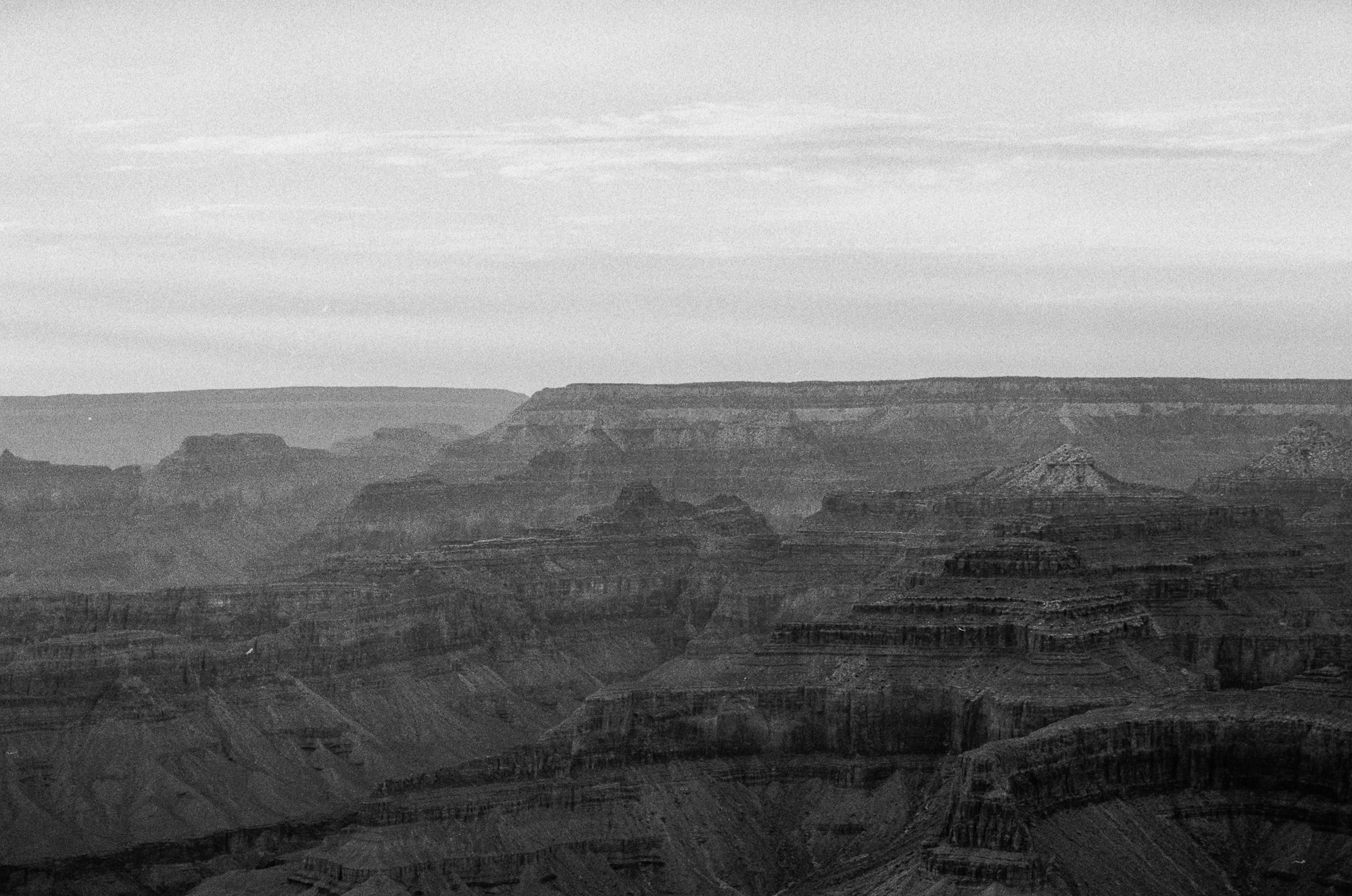 A black and white view across the Grand Canyon, with dark layered rock terraces and isolated buttes receding into haze toward a flat distant rim. The upper half of the frame is a pale, lightly clouded sky above the horizon line.