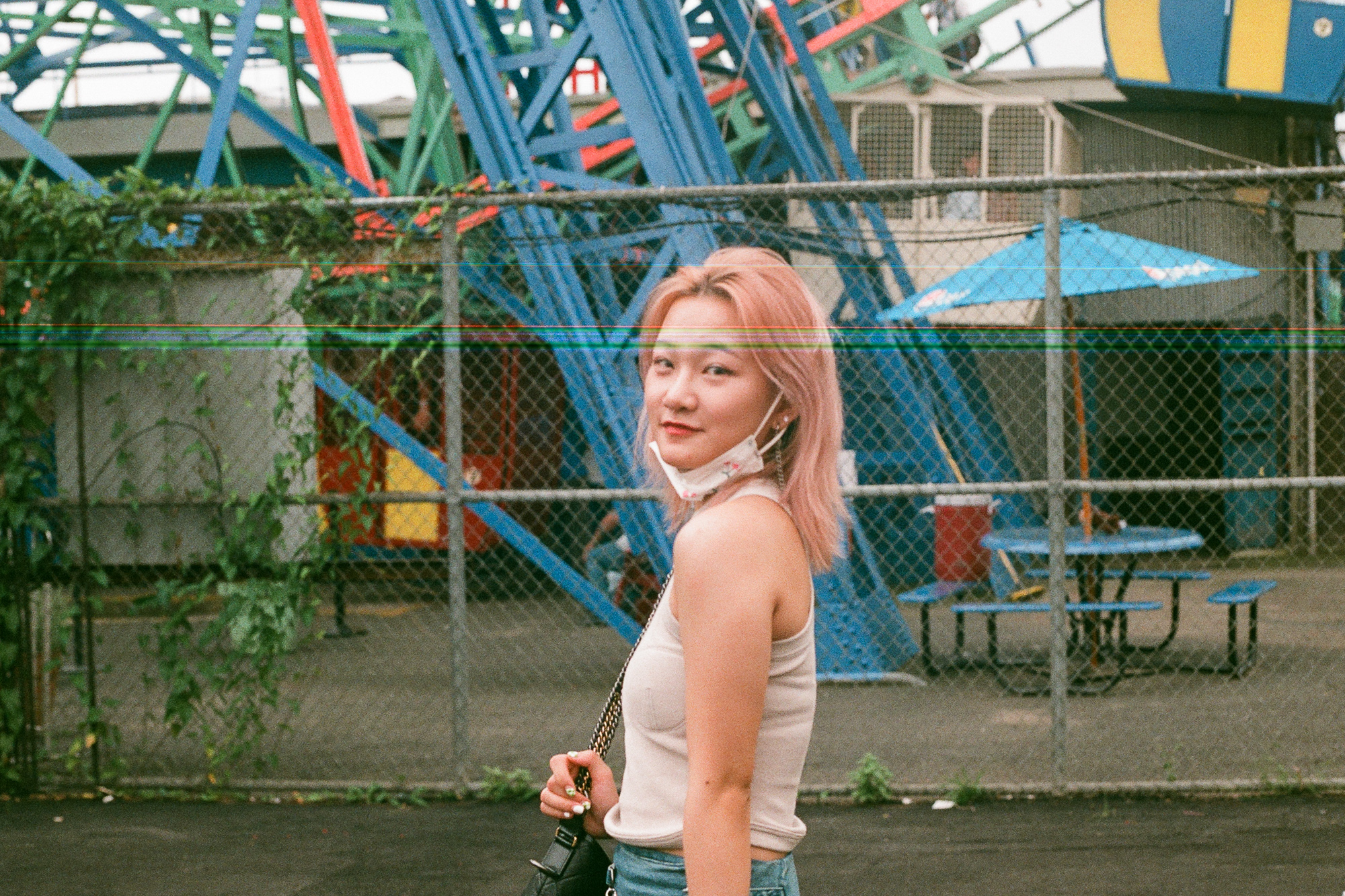 A young woman with shoulder-length pink hair in a cream ribbed tank top turns her head over her shoulder to look toward the camera, a floral mask pulled down around her neck and a chain-strap bag on her shoulder. Behind her a chain-link fence separates the walkway from blue, green and red amusement ride girders, a blue patio umbrella and empty picnic tables.