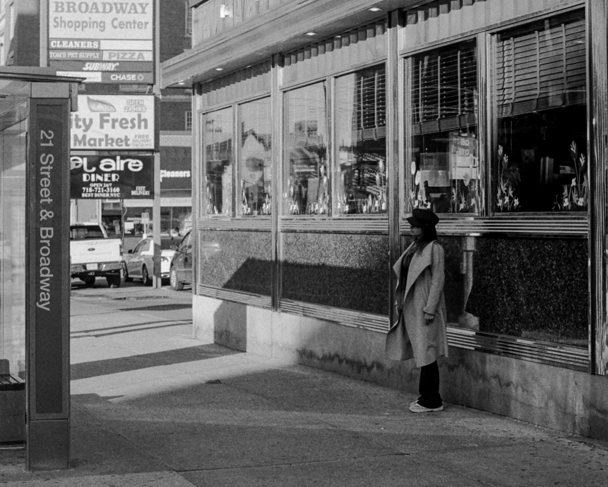 Black and white street photograph of a woman in a long light wool coat, black cap and sunglasses standing against the mirrored glass facade of a diner on a sunlit sidewalk. To the left, a bus shelter panel reads 21 Street & Broadway, with signs above for Broadway Shopping Center, City Fresh Market and El Aire Diner.