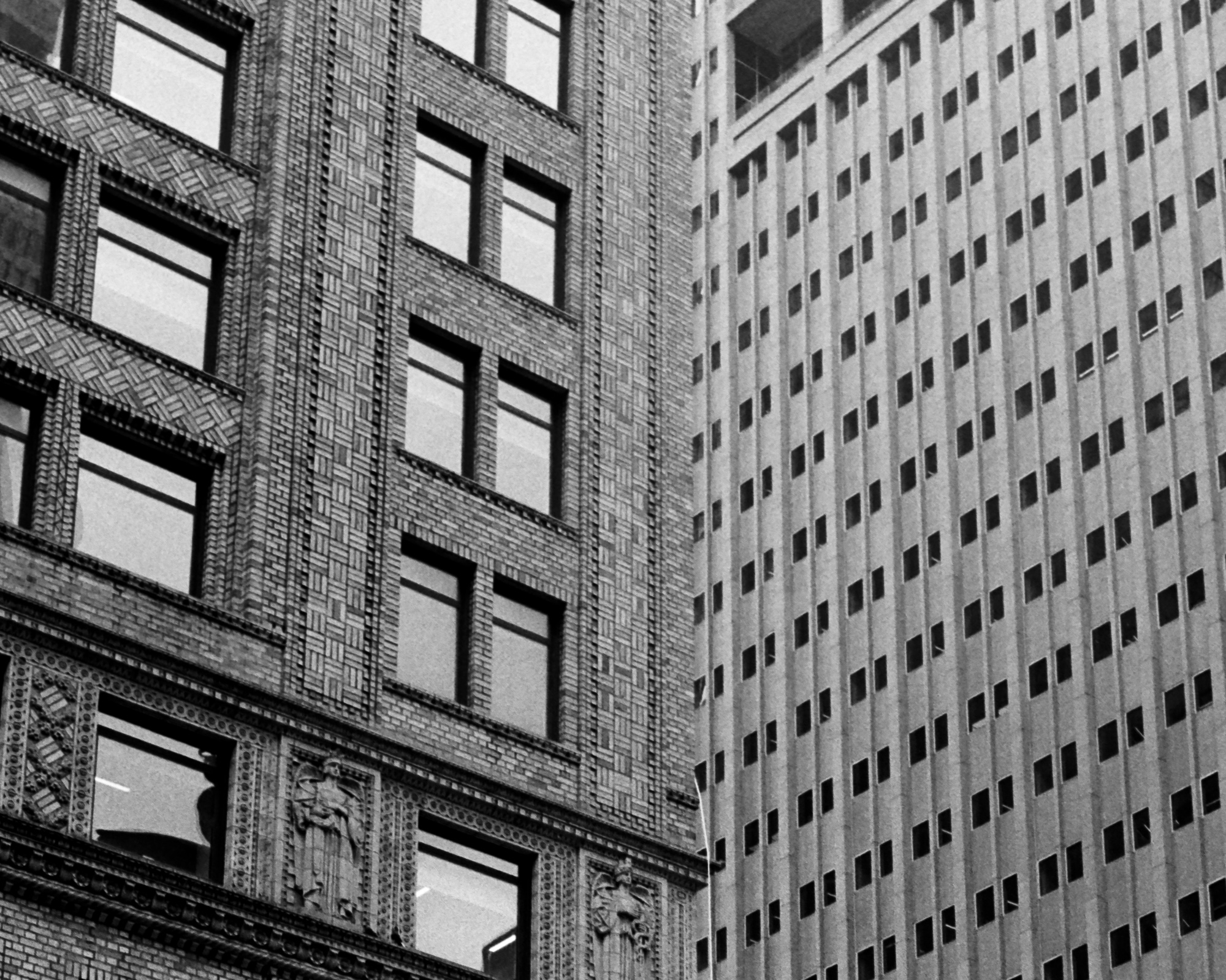 Black and white photograph of two adjoining Manhattan high-rise facades: on the left, an ornate early twentieth century building with basketweave brickwork, deep-set windows and carved robed figures beneath a cornice; on the right, a plain limestone tower rising in vertical piers with a dense grid of small dark windows.
