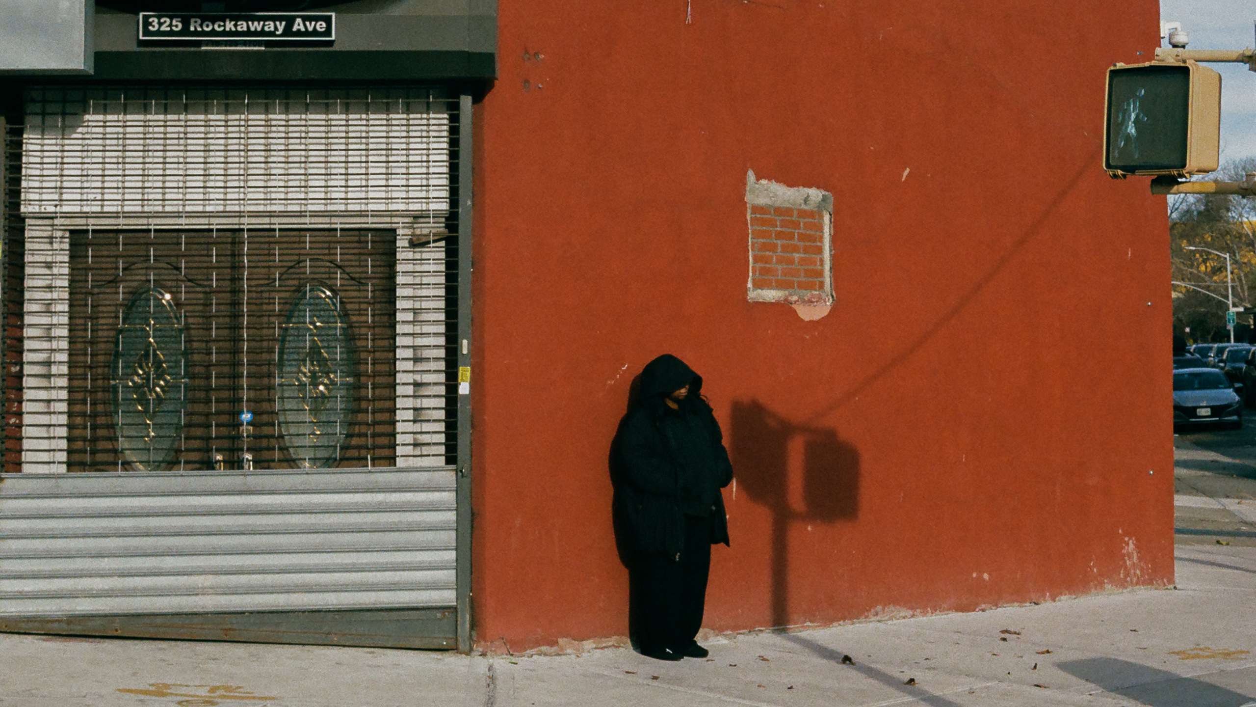 A person in a black hooded winter coat stands alone against a large red-painted corner wall in Brooklyn, head lowered, beside a patched square of exposed brick. To the left a shuttered storefront sits under a sign reading 325 Rockaway Ave, and a pedestrian signal box hangs at the right edge of the frame.