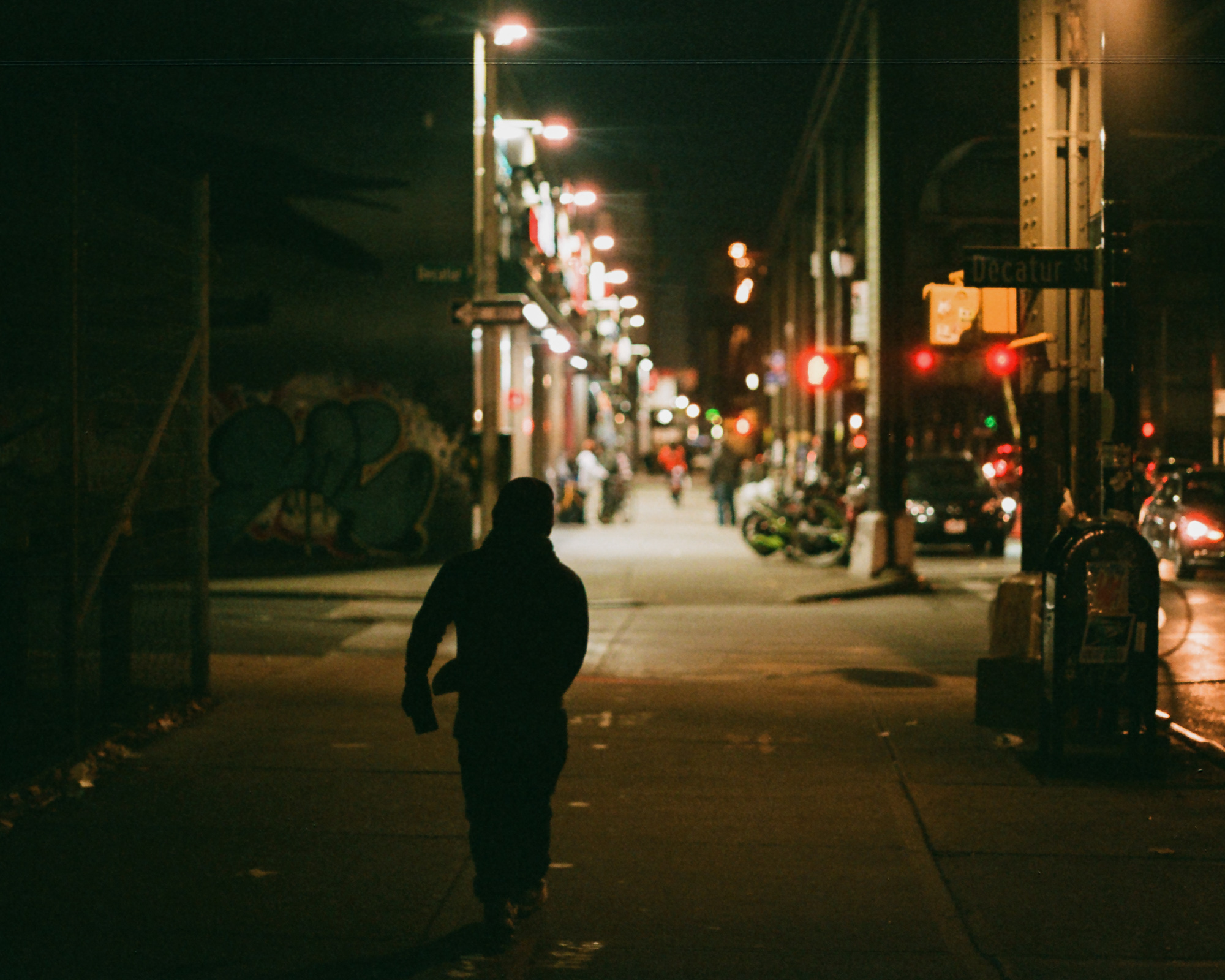 A silhouetted person in a hooded jacket walks away from the camera down a Brooklyn sidewalk after midnight, framed by a Decatur Street sign and a red traffic signal on the right. Storefront lights, parked bicycles and a graffiti-covered wall blur into the distance under an elevated train structure.