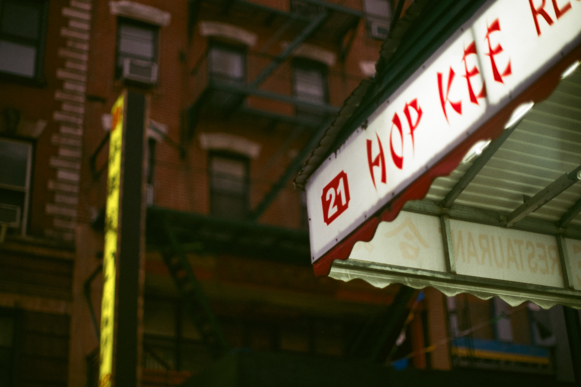 A lit white awning sign reading HOP KEE RE in red brushstroke lettering, with the street number 21 in a red box, projecting from a Chinatown storefront at night. Behind it a brick tenement facade with window air conditioners and a fire escape recedes into darkness.