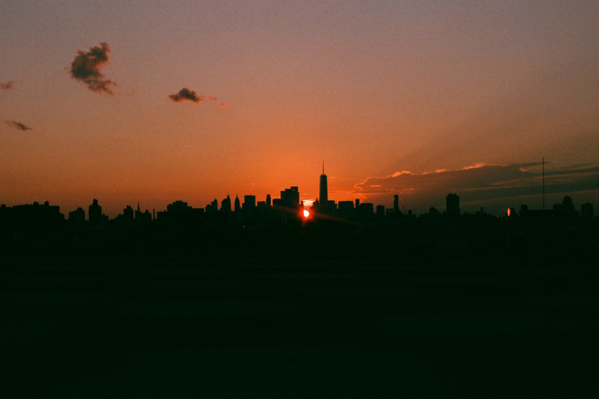 A low, flat horizon of dark building silhouettes under a deep orange and mauve sky, with a small red sun disc sinking just below the rooftops beside the spire of One World Trade Center. A few thin clouds catch pink light at upper left and along the right edge.