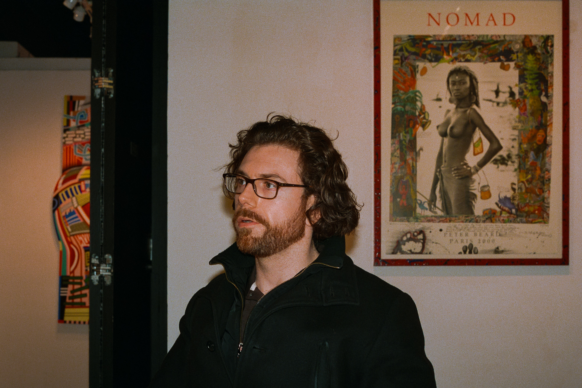 A man with shoulder-length curly brown hair, a reddish beard and dark-rimmed glasses stands in a gallery wearing a black zip-up jacket with the collar up, looking off to his right. Behind him on a pale wall hangs a framed Peter Beard 'Nomad' poster showing a standing figure surrounded by a painted collage border, with a brightly colored abstract canvas visible on the far left.