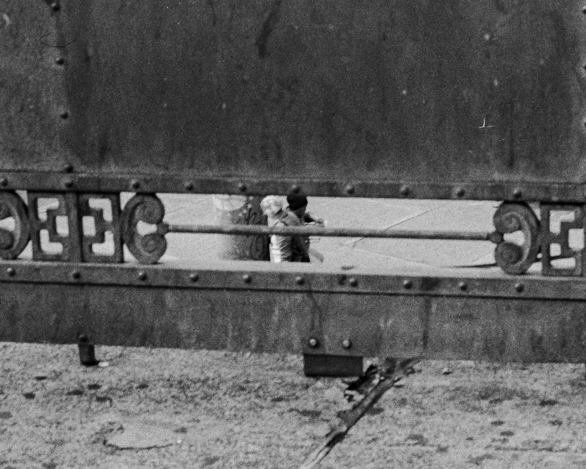 A high-contrast black and white photograph shot through the scrollwork of a riveted steel bridge railing, where two people stand close together in an embrace on the far side. One wears a dark knit cap, the other has light-colored hair, and both are framed tightly between the horizontal rail and the cast iron pattern, with gravel and steel plate filling the rest of the frame.