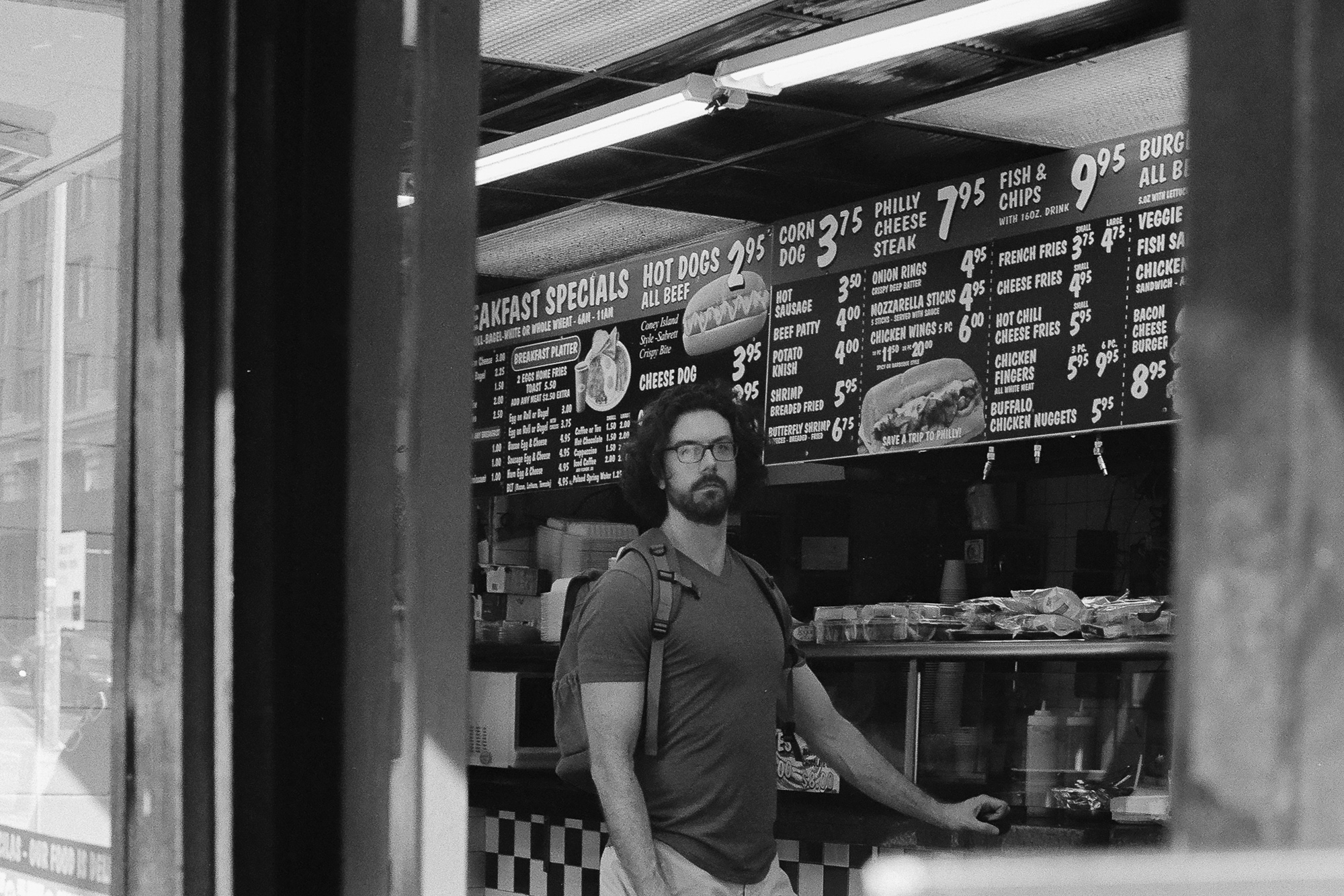 Black and white photograph of a bearded man with curly hair, glasses and a backpack standing at a fast food counter, looking off to his right. Behind him a large illuminated menu board lists hot dogs, corn dogs, Philly cheese steak, fish and chips and breakfast specials with prices.