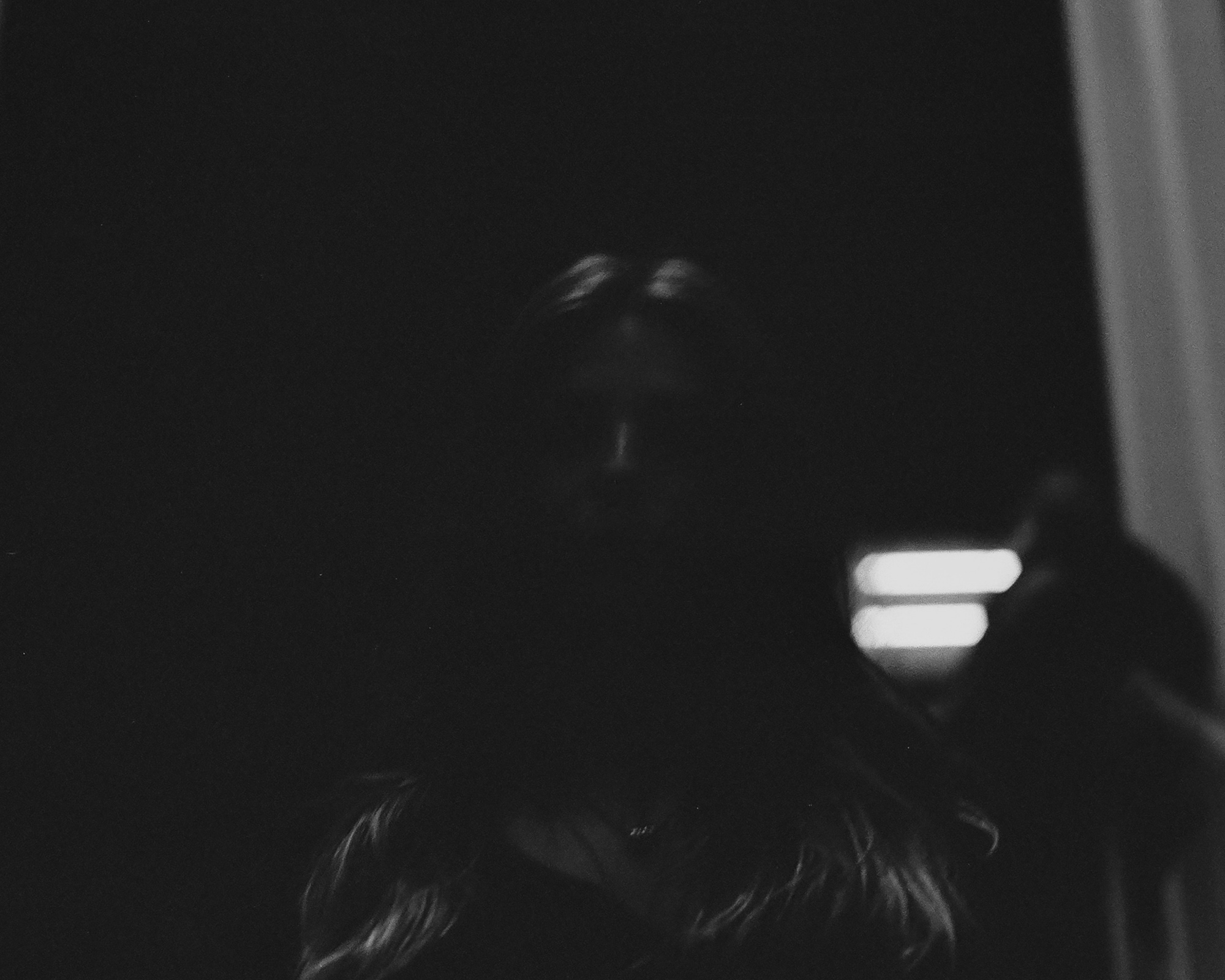 A high-contrast black and white photograph in which a person's face emerges faintly from near-total darkness, with only the brow, nose and mouth catching light. To the right, two horizontal bars of bright white light glow against the black, and long hair falls across the lower edge of the frame.
