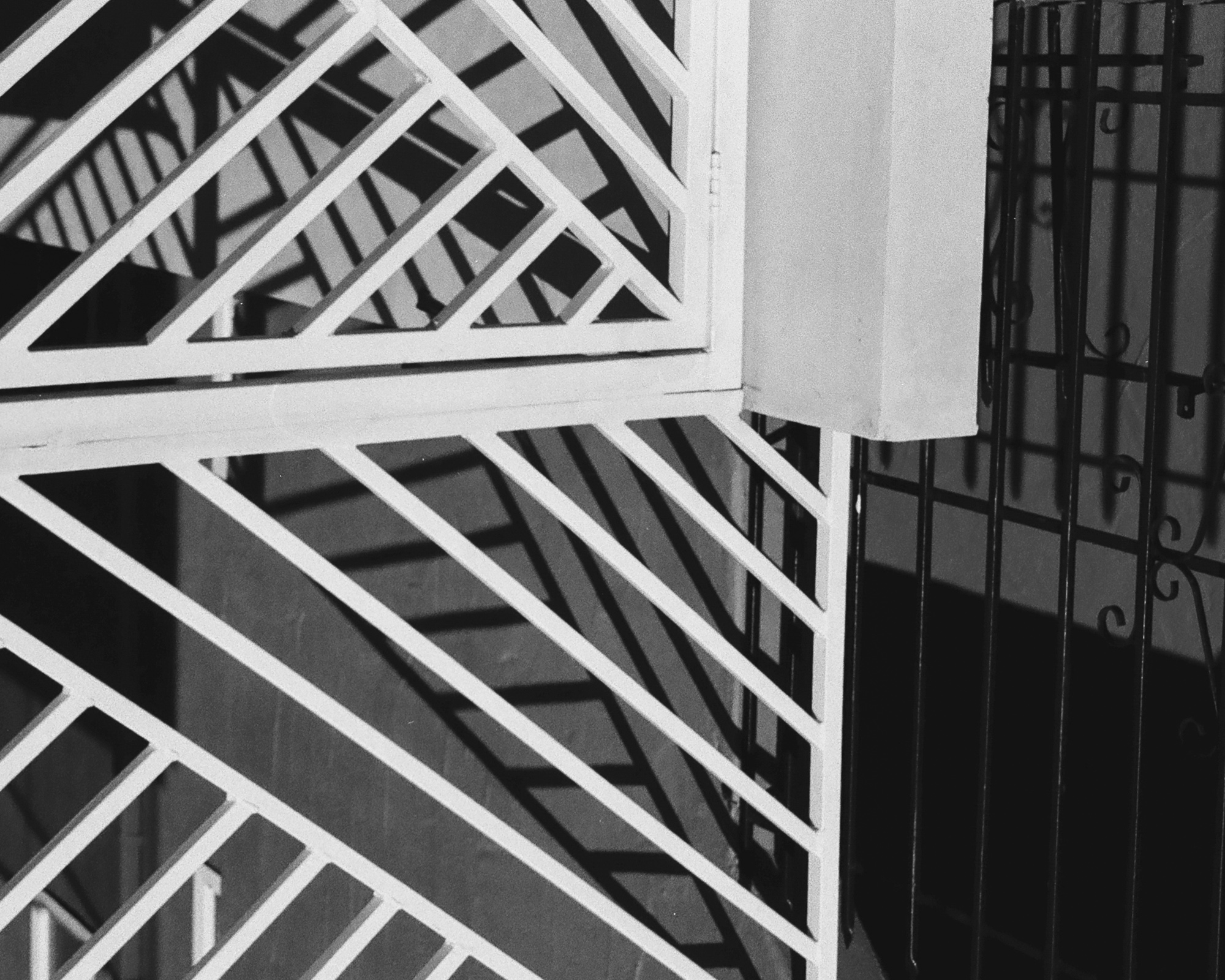 Black and white close view of a white painted metal railing with diagonal lattice bars meeting at a corner post, its shadow repeating across the wall behind it. To the right, a dark wrought iron gate with scroll details stands in deep shadow.