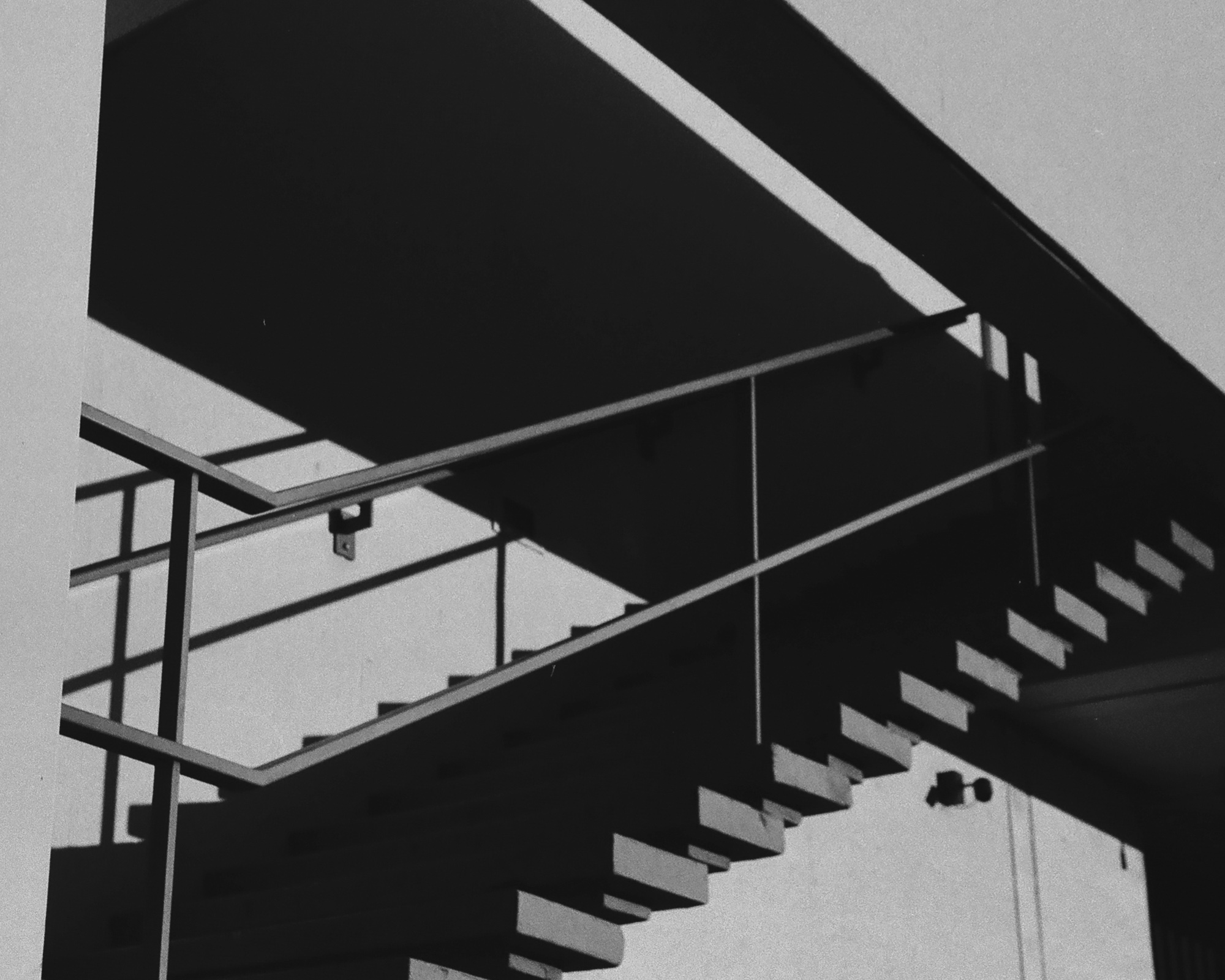 Black and white photograph looking up at an exterior concrete staircase, its open treads cantilevered from a diagonal beam and casting deep shadows on a pale stucco wall. A thin steel handrail cuts across the frame beneath the dark underside of an upper landing.