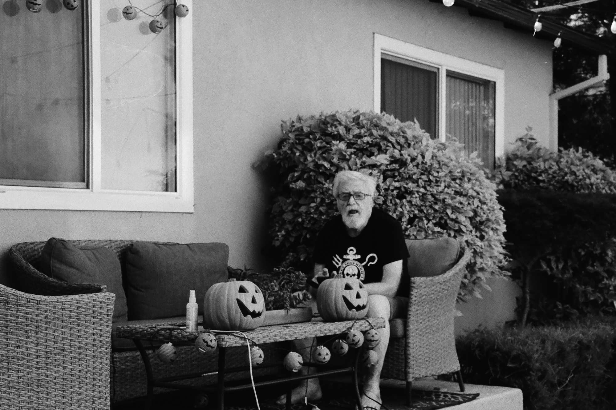 A white-haired man in glasses and a dark graphic T-shirt sits on a wicker patio chair, holding a carved jack-o'-lantern in his lap while another sits on the low table in front of him. Strings of small pumpkin-shaped lanterns drape across the table and the house wall behind, with a large leafy shrub and window frames filling the background.