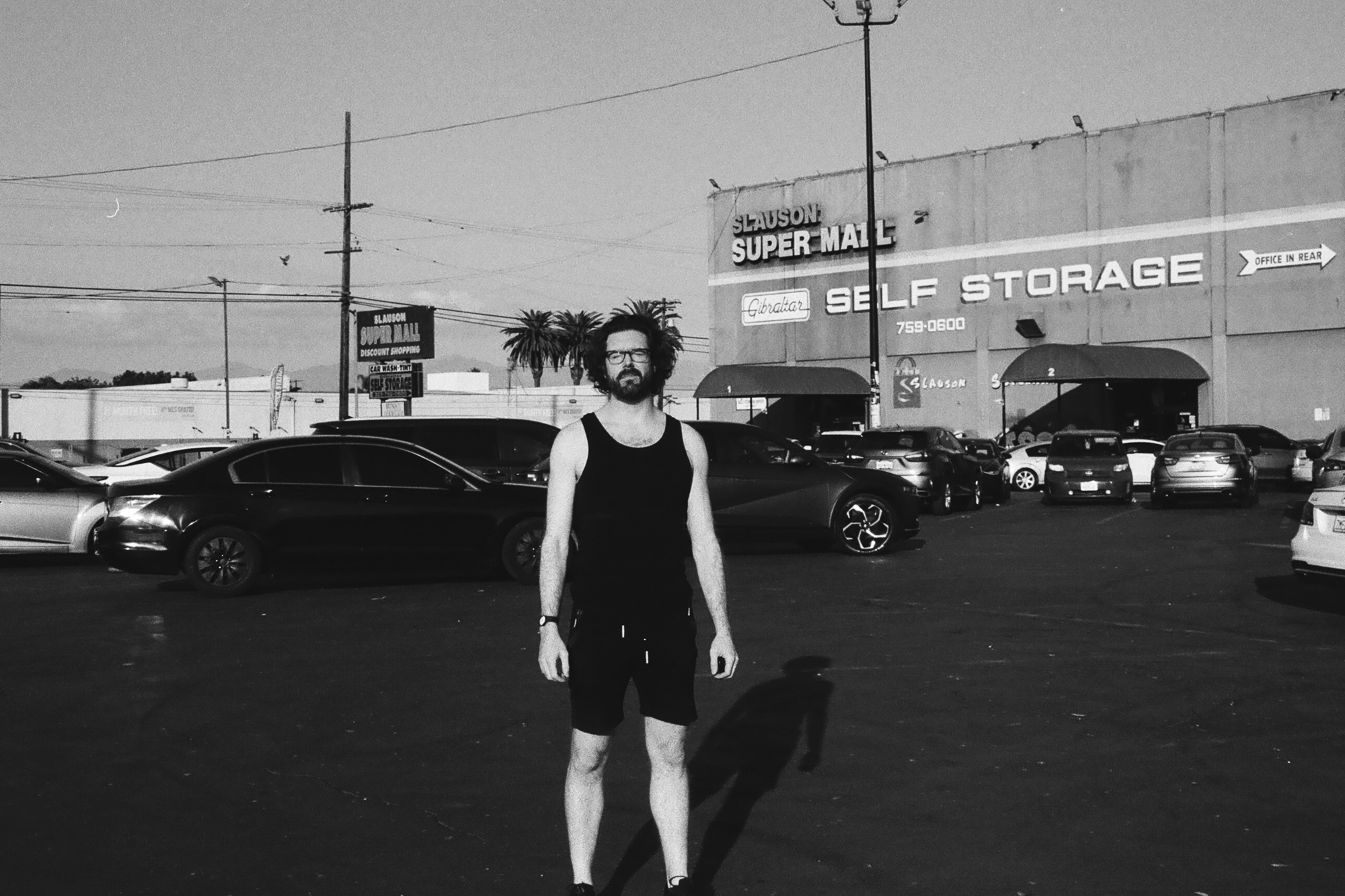 A bearded man with curly hair and glasses stands in a sunlit asphalt parking lot wearing a black tank top and shorts, his shadow stretching to the right. Behind him are parked sedans and SUVs and a long concrete building painted with signs reading Slauson Super Mall and Gibraltar Self Storage, with palm trees and power lines against a pale sky.