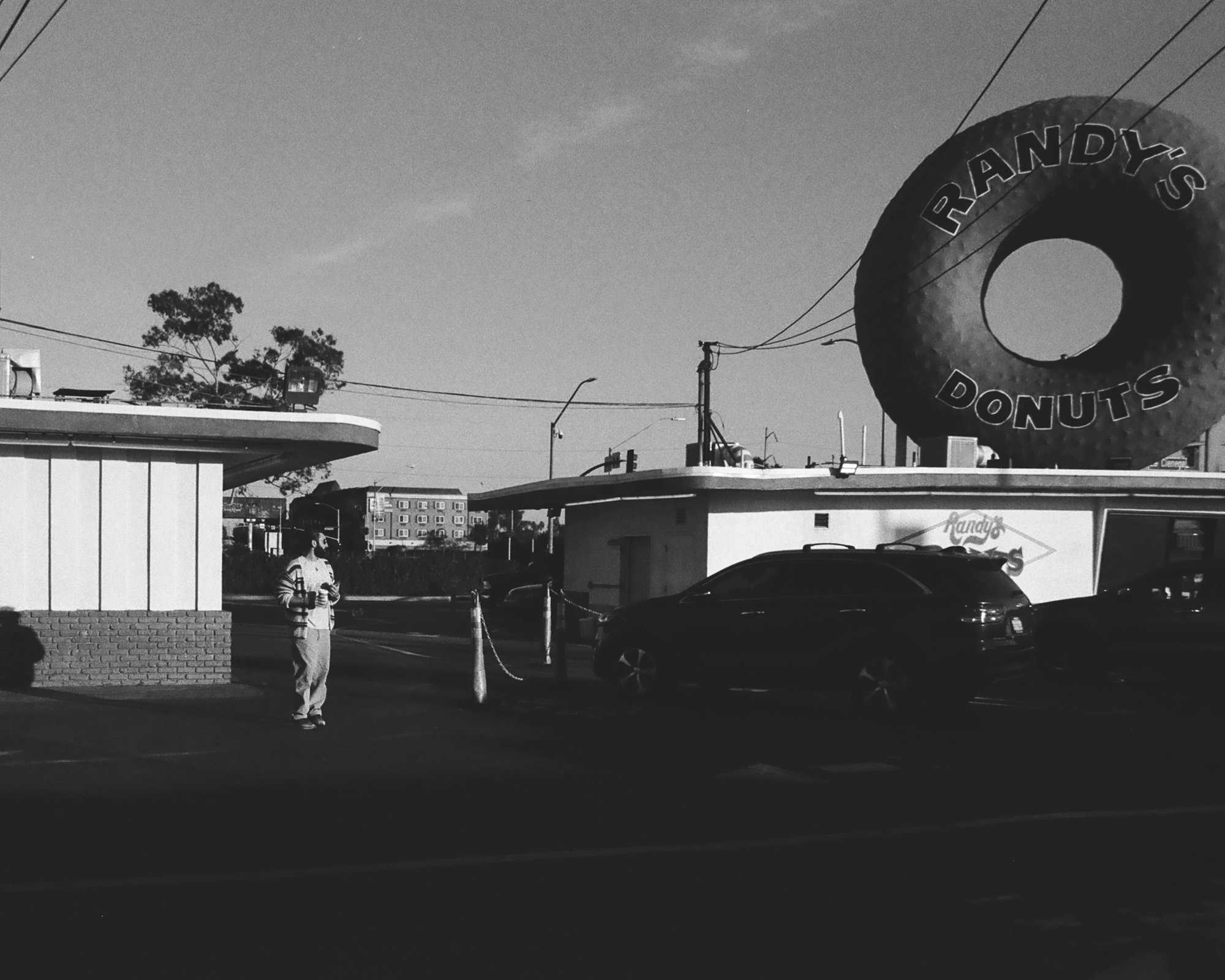 Black and white photograph of Randy's Donuts in Los Angeles, with the enormous rooftop donut sign rising above a low white building and a dark SUV parked in the drive-through lane. A bearded man in a light hoodie and sweatpants stands in sunlight at the edge of the lot holding a coffee cup, with power lines crossing the pale sky above.
