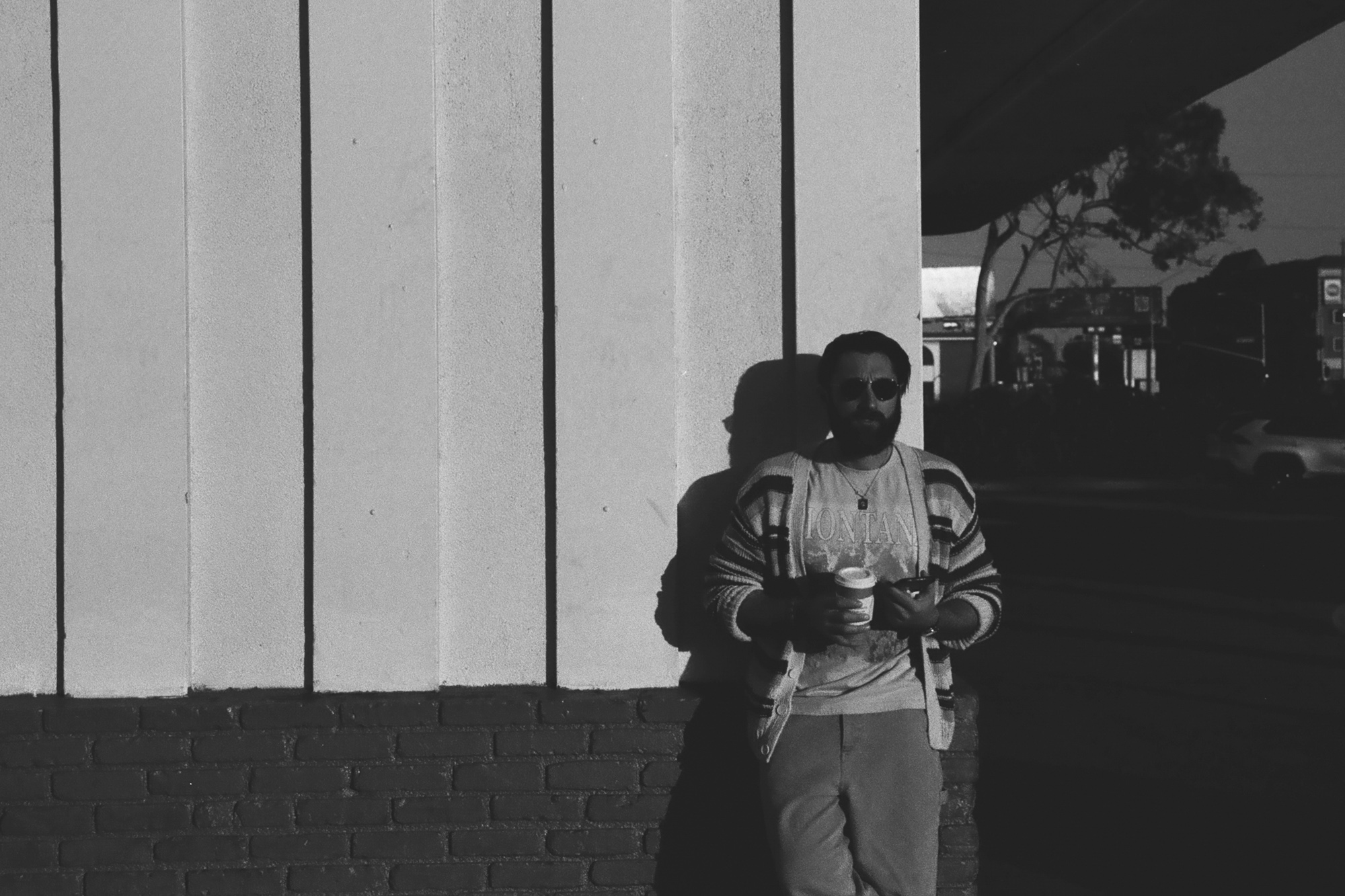 Black and white photograph of a bearded man in dark sunglasses, a striped cardigan and a graphic tee reading MONTANA, standing against a tall ribbed white concrete wall with a brick base while holding a paper coffee cup. His shadow falls sharply on the wall behind him, and the right half of the frame drops into deep shade where a tree, a distant car and storefront signs are barely visible.