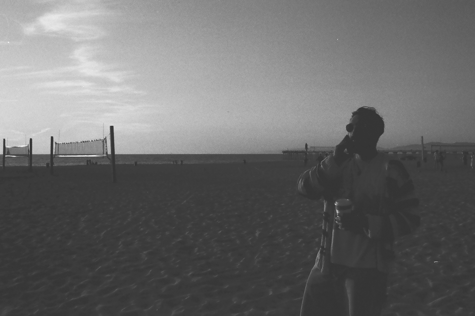 A black and white photograph of a person standing in silhouette at the right edge of a wide sandy beach, wearing sunglasses and a knit sweater, holding a lidded coffee cup and a bag while raising one hand toward their face. In the distance are two volleyball nets on poles, the flat ocean horizon, and a pier extending into the water under a pale hazy sky.