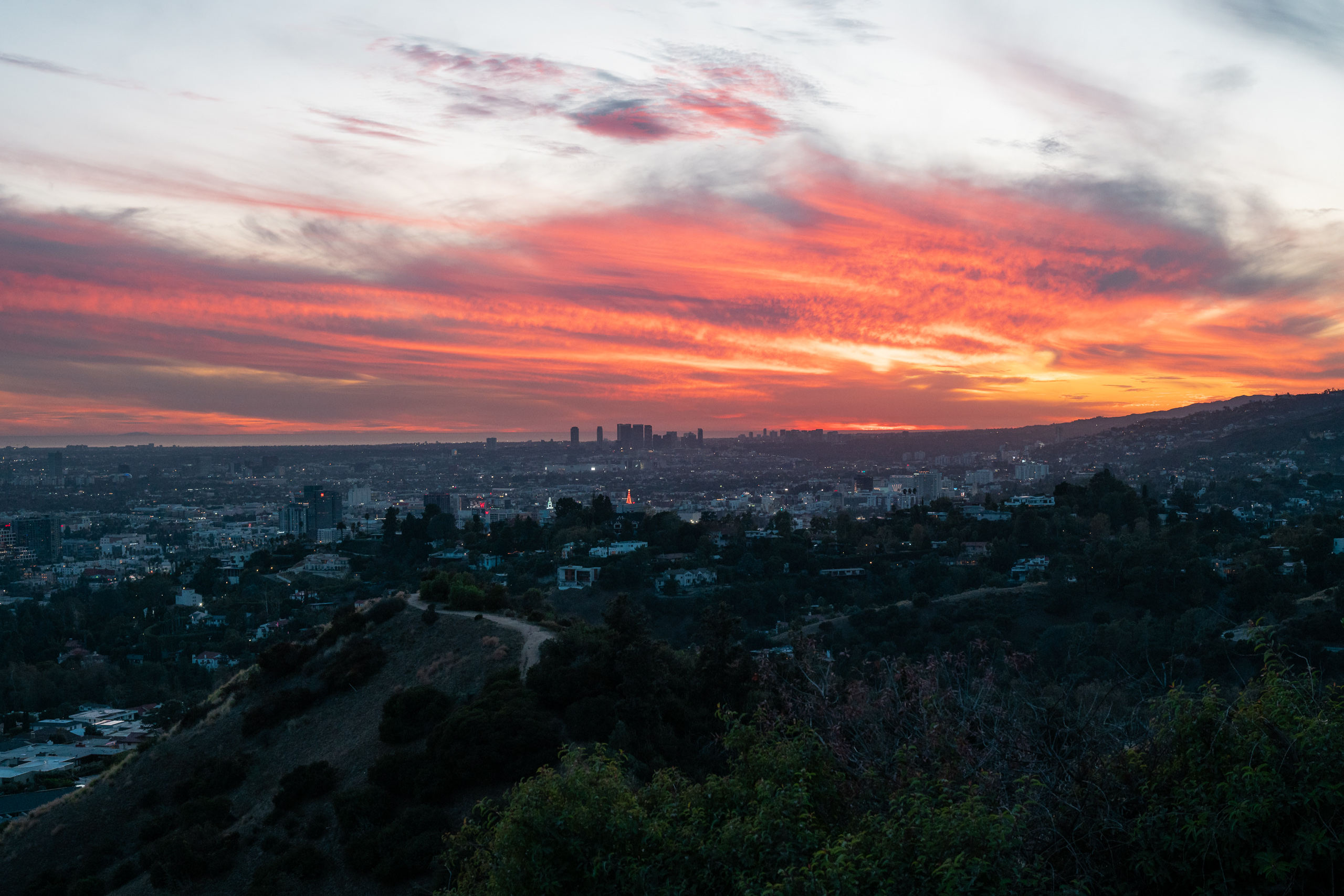 A wide view from a dark chaparral hillside looking west over Los Angeles at dusk, with a dirt footpath winding along the ridge and city lights spreading across the basin. The sky is streaked with bands of red, orange and grey cloud above the distant Century City towers and a hazy line of ocean on the horizon.