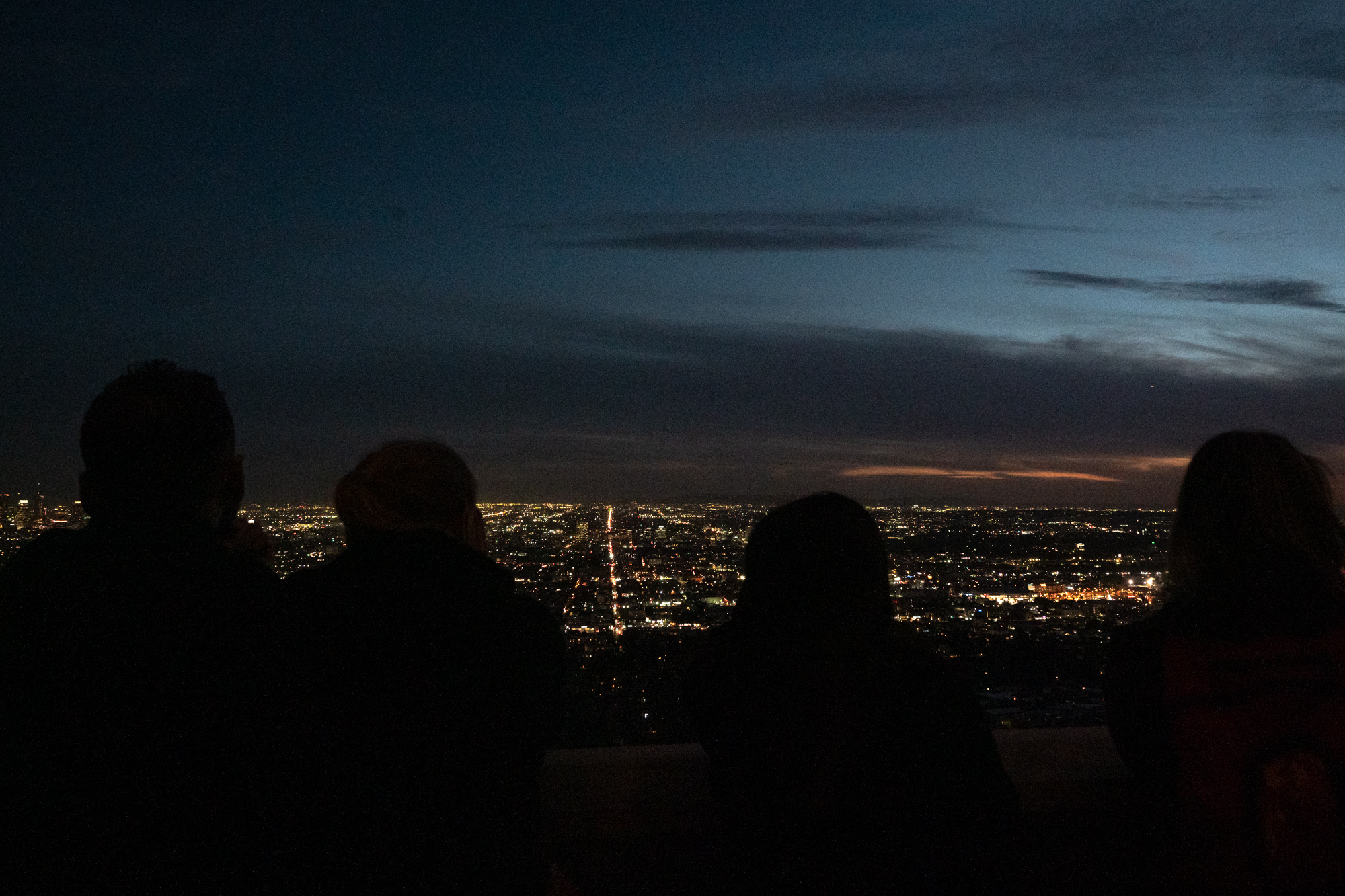 Four people appear as dark silhouettes in the foreground, looking out over a vast grid of Los Angeles city lights from an elevated overlook. Above the glowing horizon, layered clouds streak a deep blue twilight sky with a thin band of orange near the skyline.
