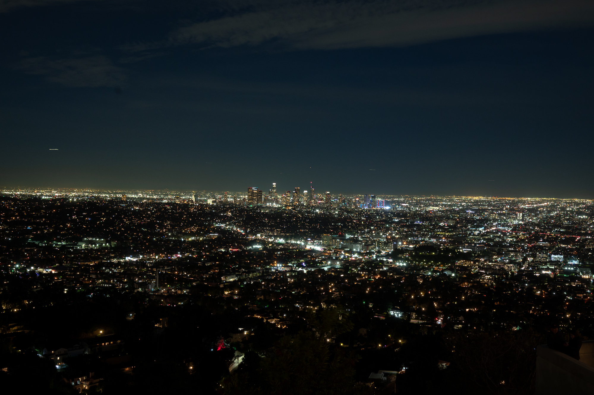 A nighttime view across Los Angeles from an elevated hillside, with thousands of scattered street and house lights spreading toward the horizon and the clustered towers of the downtown skyline glowing at center. The upper half of the frame is dark blue sky streaked with a thin band of cloud.