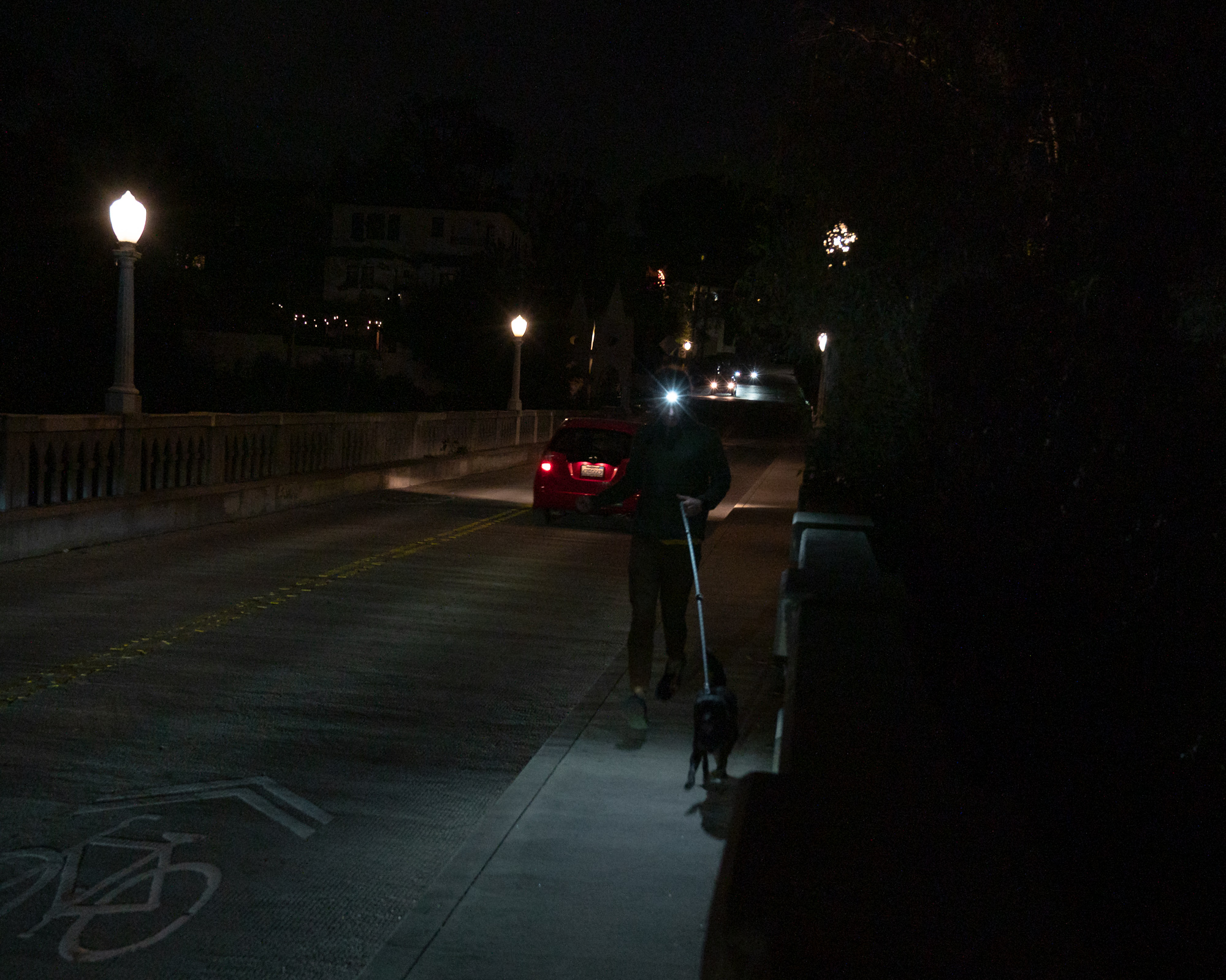 At night, a person wearing a bright headlamp walks a black dog on a leash along the sidewalk of a concrete balustrade bridge, passing a parked red hatchback with its brake lights glowing. Ornate street lamps and distant headlights recede up the road toward dark hillside houses.