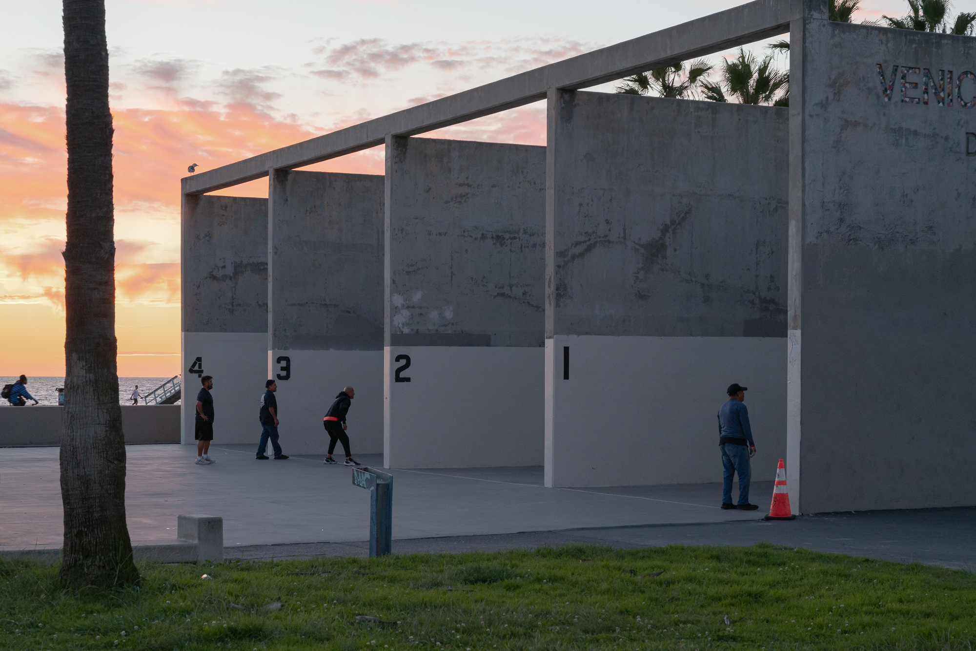Four numbered concrete handball courts at Venice Beach stand against a pink and orange sunset sky, with players walking between the open bays and a man in a cap and blue jacket standing near an orange traffic cone at right. A palm tree trunk fills the left edge of the frame and the ocean is visible in the gap beyond the courts.