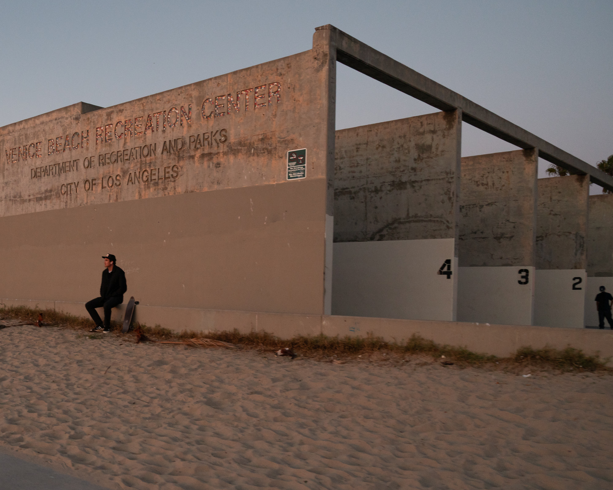 A young man in a black cap, jacket and jeans leans against the low concrete wall of the Venice Beach Recreation Center at dusk, his skateboard propped upright beside him. The weathered wall carries raised lettering reading Venice Beach Recreation Center, Department of Recreation and Parks, City of Los Angeles, with numbered handball courts receding to the right and a second figure standing in the distance.