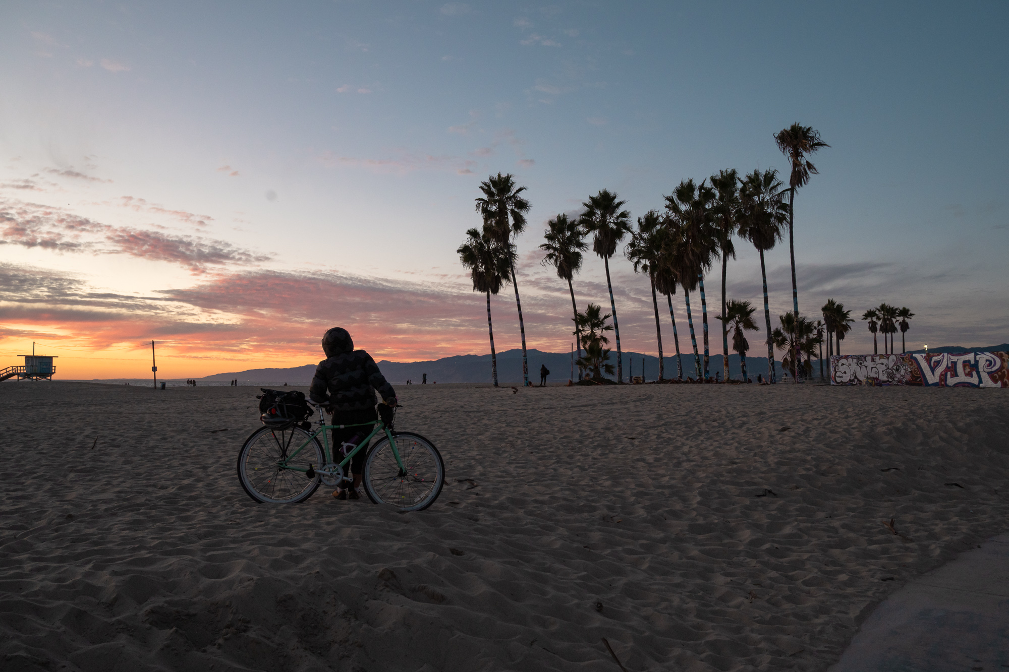 A person in a helmet and dark puffy jacket stands beside a mint green bicycle with a loaded rear rack on wide beach sand, facing a row of tall palm trees silhouetted against an orange and pink sunset sky. A lifeguard tower sits at far left, distant mountains line the horizon, and a graffiti-covered wall runs along the right edge.