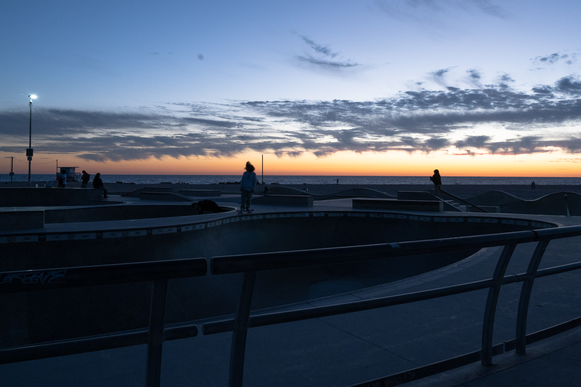 A skateboarder with wind-blown hair stands in silhouette on the lip of a concrete bowl at a beachfront skatepark, backlit by an orange band of light along the ocean horizon. Metal handrails cut across the darkened foreground while other figures and a lifeguard tower sit along the far edge of the park.