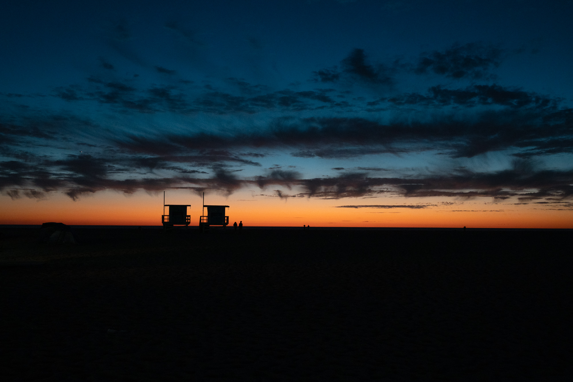 Two lifeguard towers stand in silhouette on a darkened beach beneath a band of orange afterglow along the horizon, with streaked blue clouds filling the upper sky. Two small figures stand on the sand to the right of the towers, with more distant walkers further along the shore.