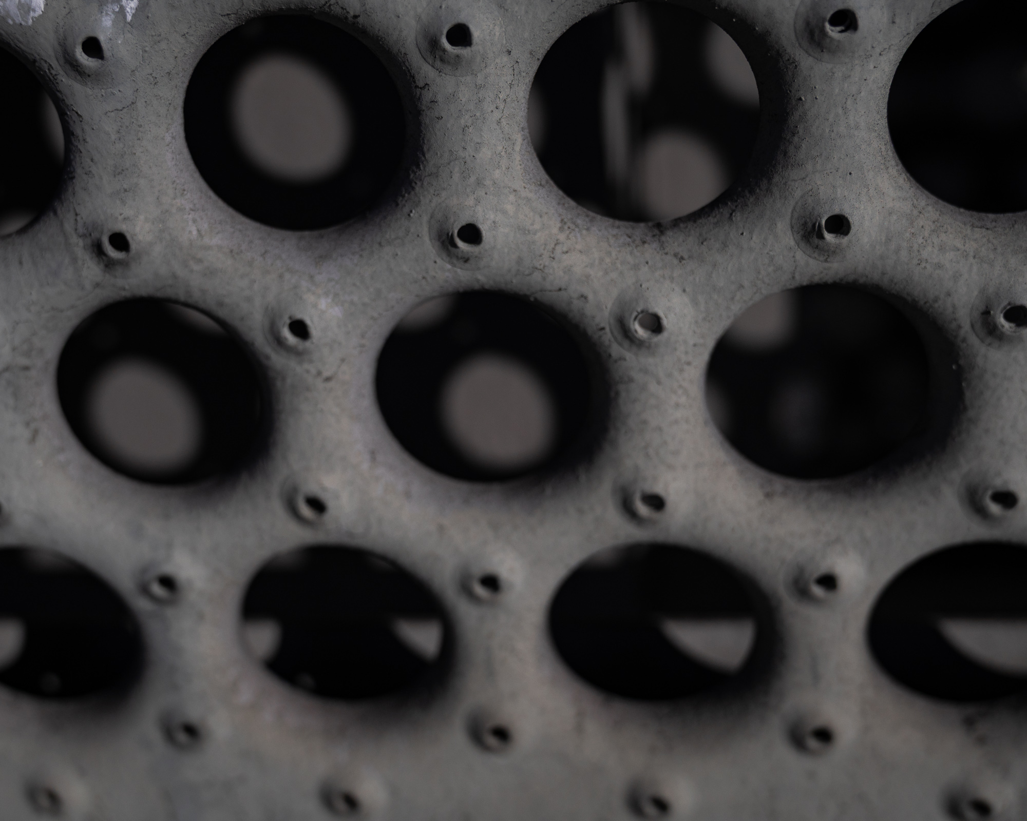 Extreme close-up of a gray, pitted metal surface punctured by rows of large dark circular holes, with small pinholes set between them; the lower holes blur into ellipses as the plate angles away from the camera.