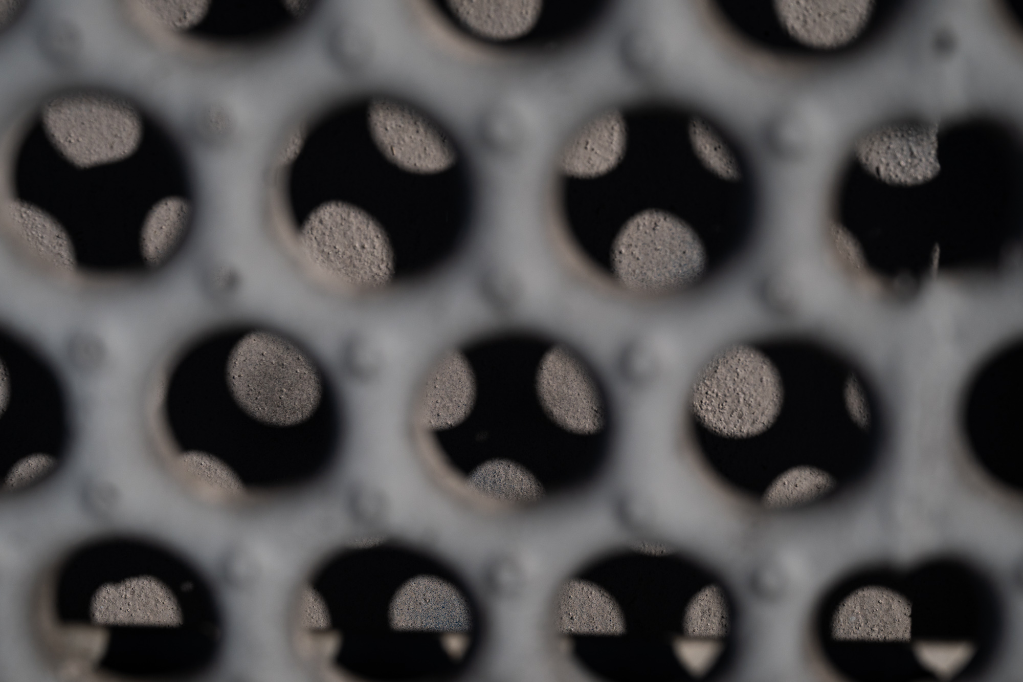 An extreme close-up of a gray perforated metal or concrete panel, its round holes arranged in an offset grid. Through each opening, black voids and pale textured shapes appear, with focus falling off sharply toward the edges of the frame.