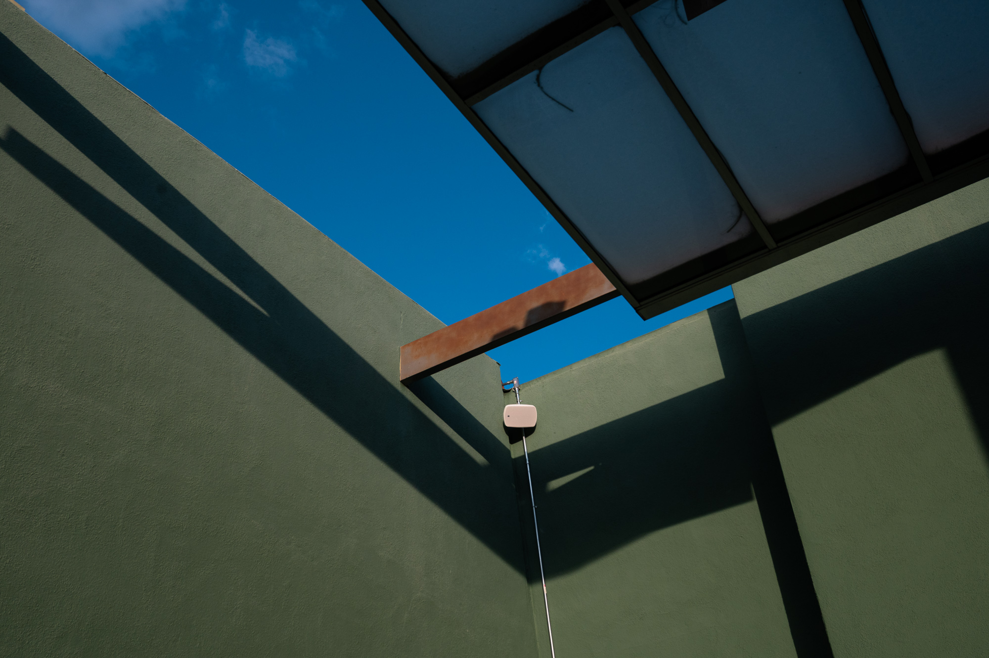 Looking up from inside a courtyard with olive green stucco walls that meet at a corner, framing a wedge of deep blue sky. A rust colored steel beam crosses the opening, a translucent paneled canopy extends from the upper right, and a small cream colored fixture with a thin pole is mounted on the far wall.