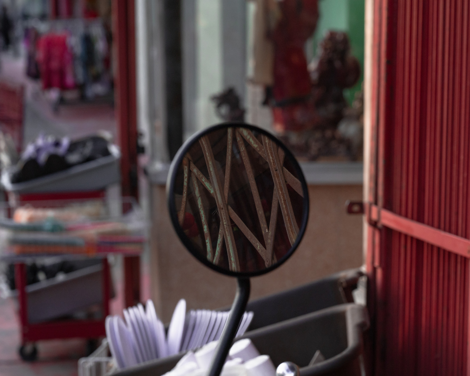 A round black scooter mirror stands in sharp focus at center frame, its glass filled with the reflection of rusted metal bars crossing at sharp diagonals. Behind it the street blurs into a red corrugated shutter, shop doorways, carts of merchandise and a bundle of white plastic cutlery in the lower foreground.