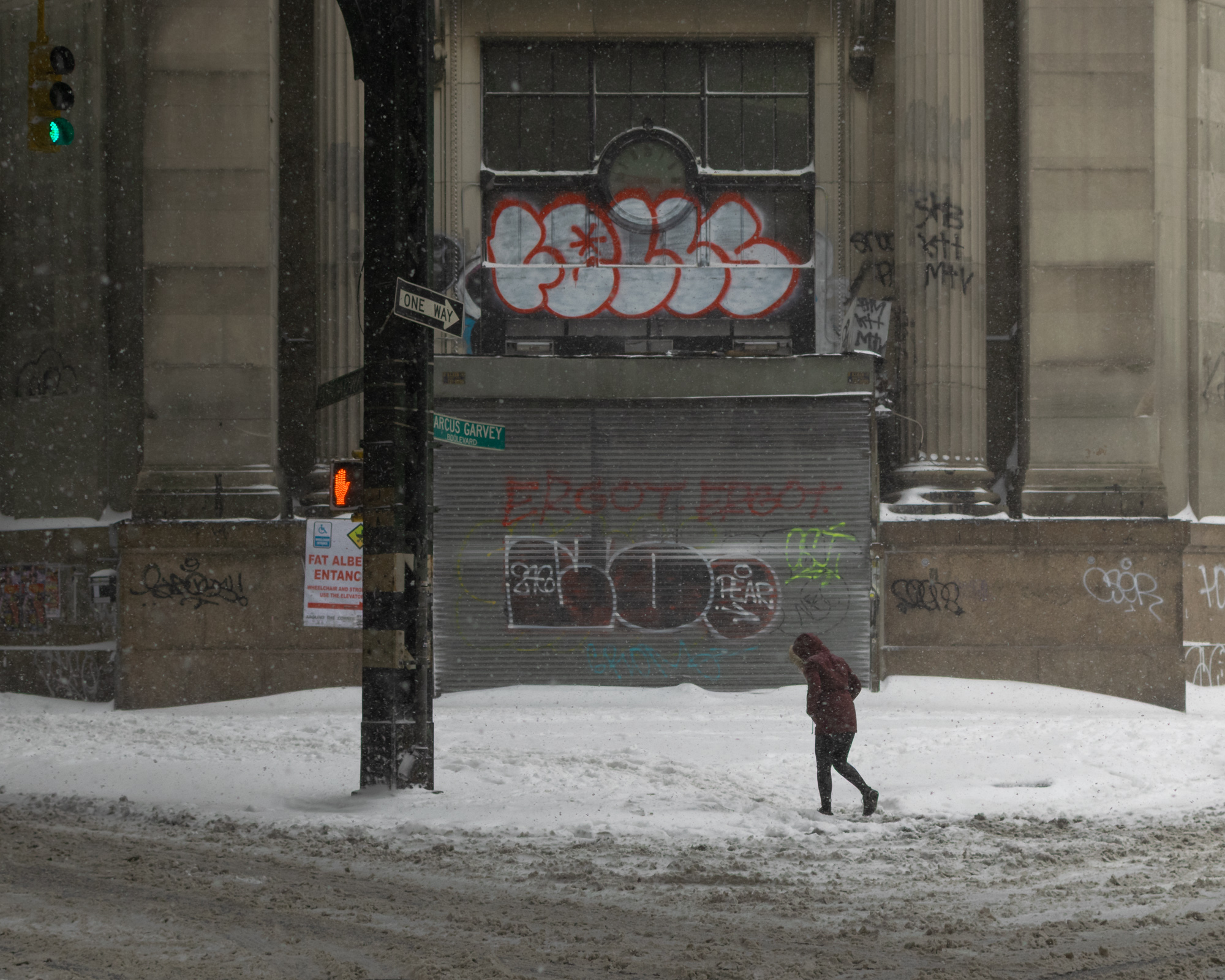 A single figure in a maroon hooded parka trudges left to right through deep unplowed snow at a street corner, head down against the storm. Behind them a shuttered storefront covered in red and silver graffiti sits under a stone facade, with a green Marcus Garvey Boulevard sign, a One Way arrow, and an orange hand crosswalk signal on the traffic pole.
