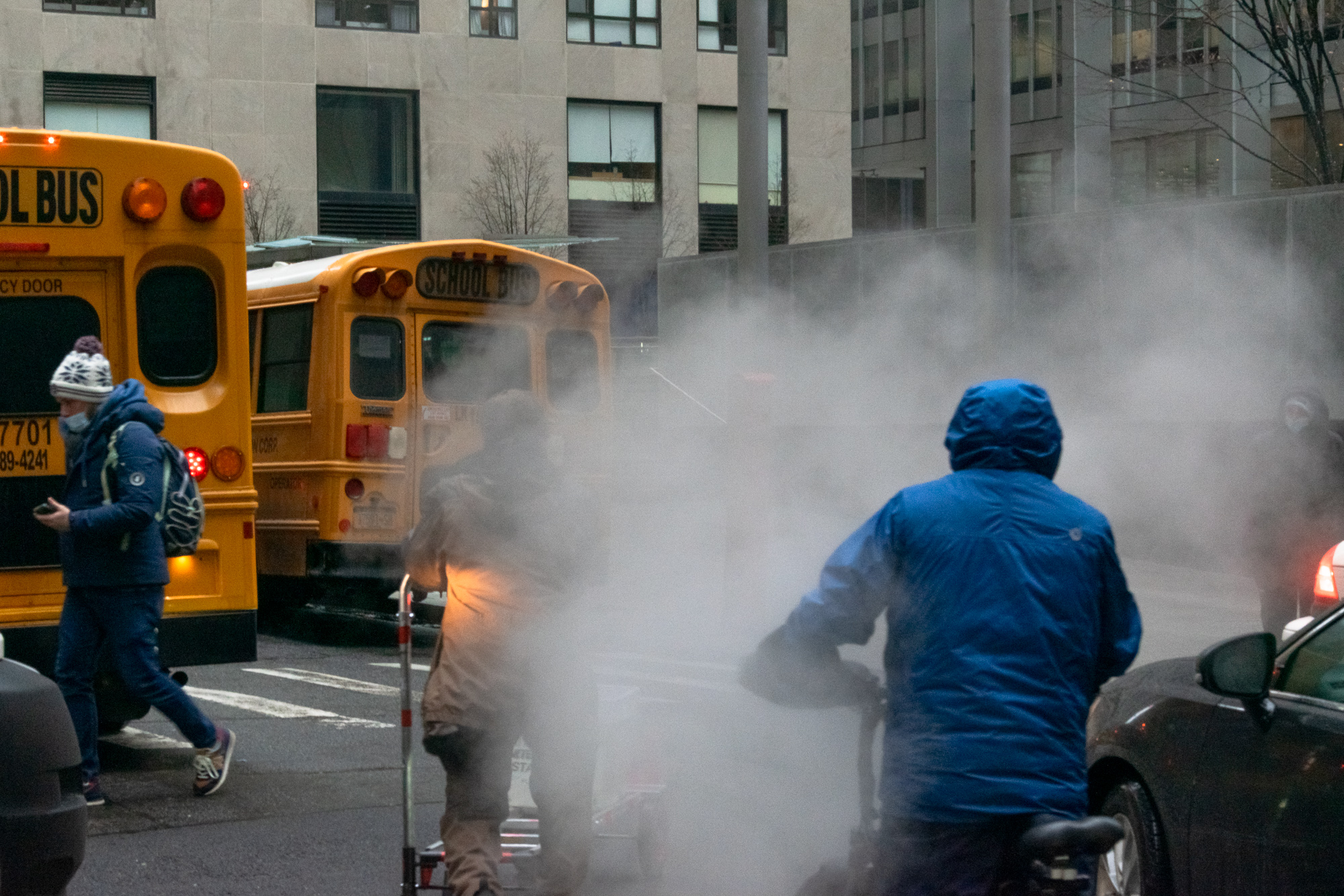 A cyclist in a blue hooded rain jacket rides through a thick cloud of white street steam on a Manhattan street, with two yellow school buses stopped at the curb behind and a pedestrian in a knit cap crossing at left.