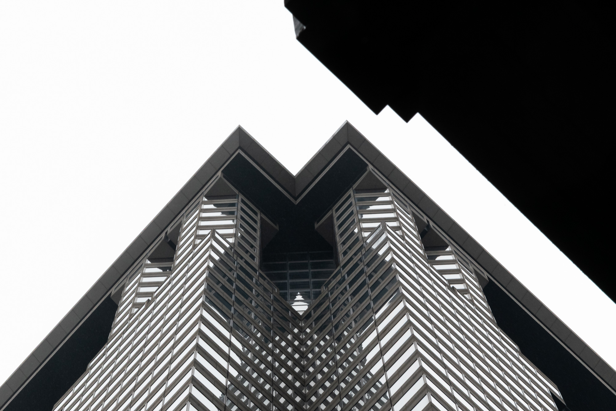 Looking straight up at the corner of a dark-clad high-rise whose banded glass and metal facade forms a chevron pattern converging on a V-shaped notch in the building's crown, set against a flat white overcast sky with a black silhouetted rooftop cutting into the upper right of the frame.