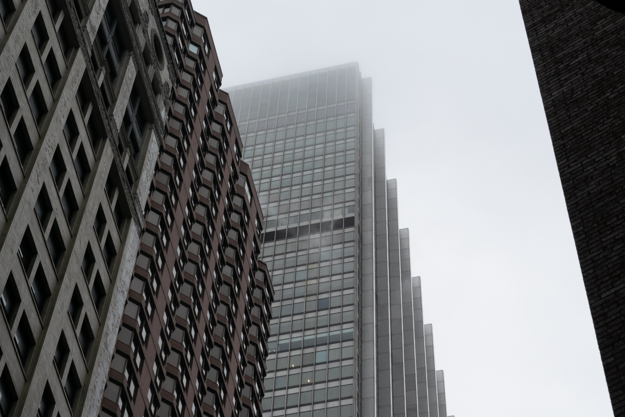 Looking up from street level at a modern glass-and-steel skyscraper whose upper floors dissolve into low gray fog, flanked on the left by a stepped brown brick tower with bay windows and a pale masonry building, and on the right by the dark edge of a brick wall.