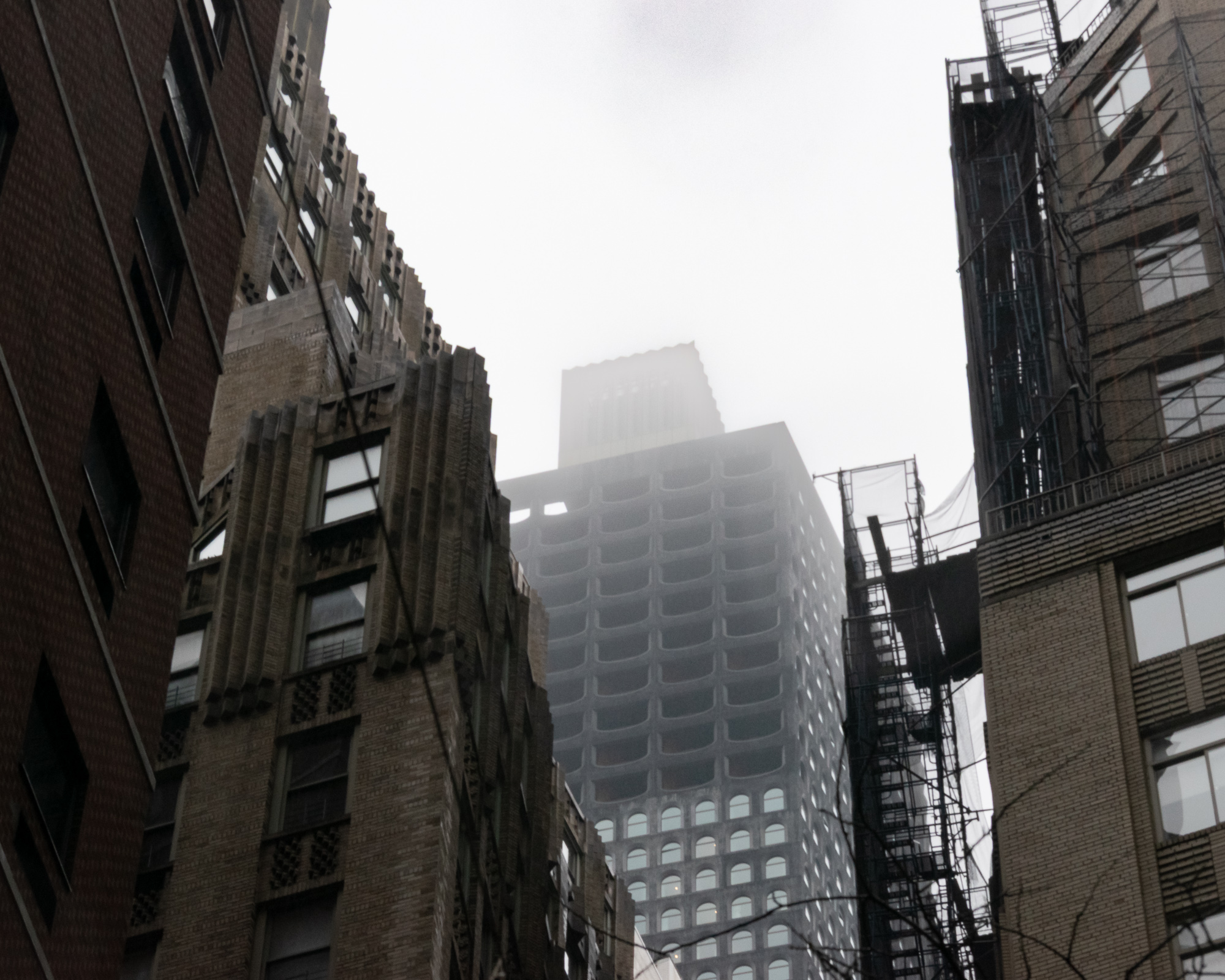 Upward view from a narrow Manhattan street: a modernist tower with rows of rounded concrete window bays rises into thick fog, its stone crown barely visible at the top. An ornate Art Deco brick facade with stepped buttresses fills the left frame, and a scaffold-wrapped building with fire escapes and white netting stands at right.