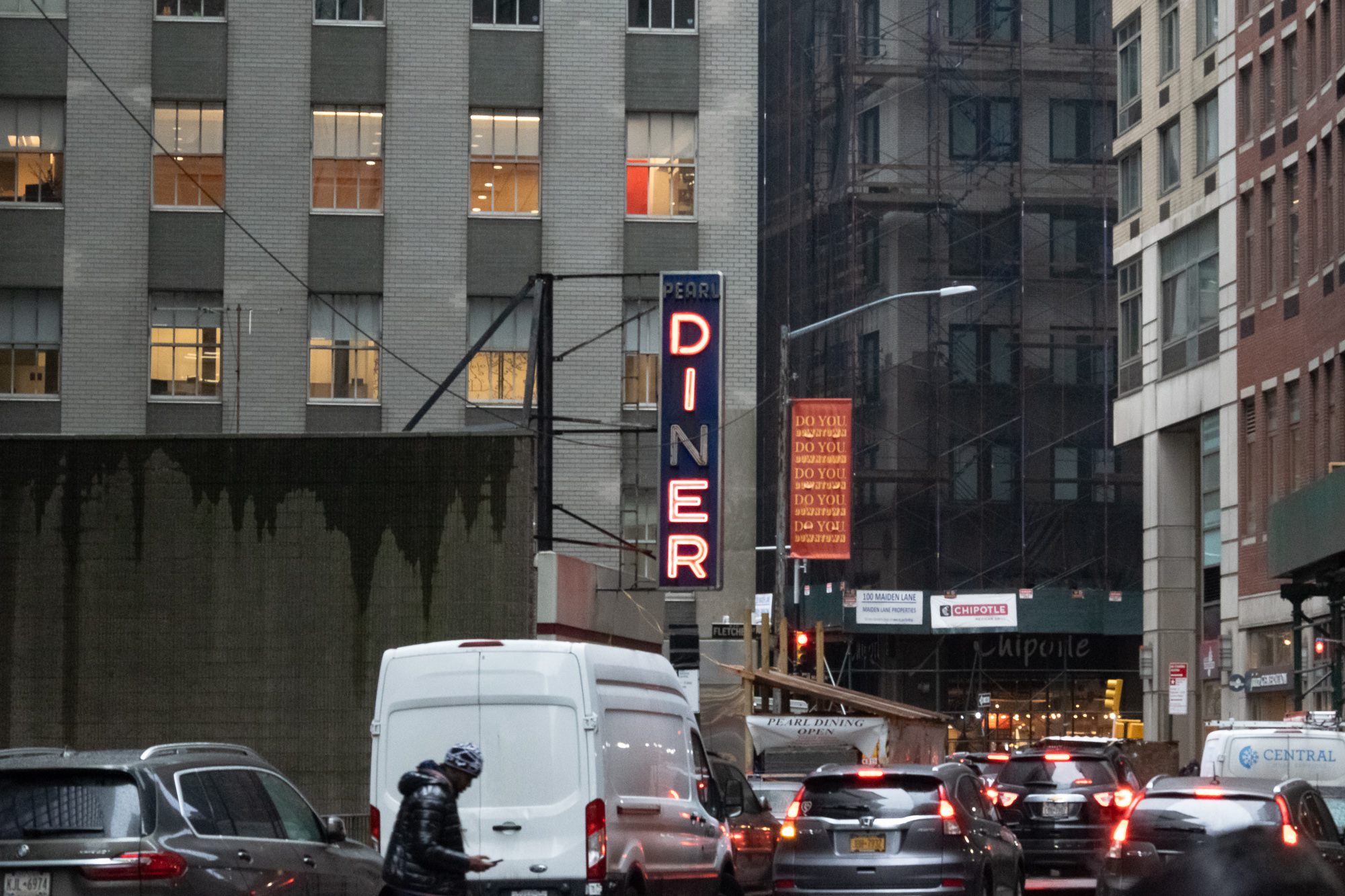 A tall vertical neon sign reading PEARL DINER glows red and blue on the side of a gray brick building in Lower Manhattan, with scaffolding, orange banners and a Chipotle storefront beyond it. Below, a man in a black puffer jacket and patterned hat walks past a white cargo van amid a line of cars with brake lights on.