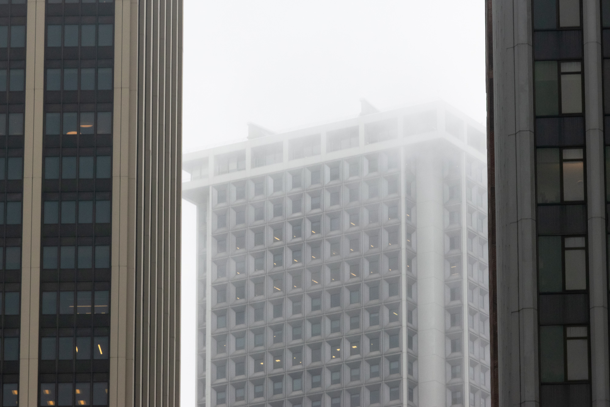 Seen through a narrow gap between two dark high-rise facades, a pale concrete tower with deep recessed square windows rises into low fog that erases its upper corner. A few interior lights glow yellow behind scattered windows.