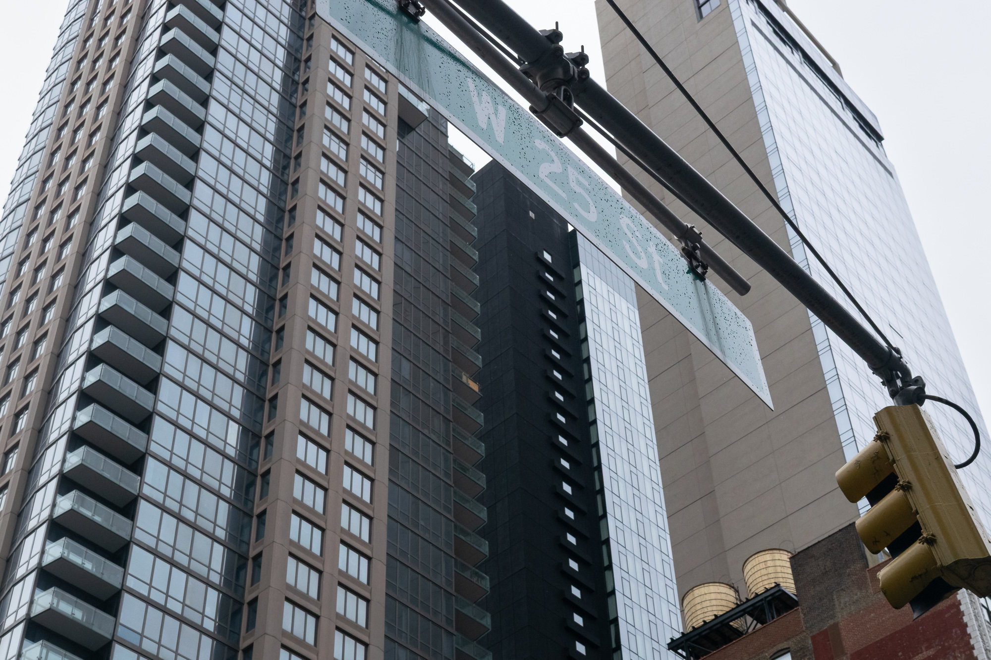 Looking up at the back of a green W 25 St street sign mounted on a black mast arm, its lettering reversed and visible through the sign, with glass and brick residential towers and a yellow traffic signal filling the frame.