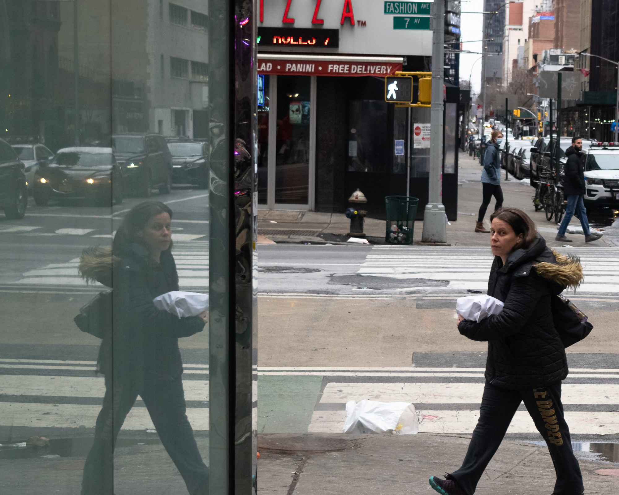 A woman in a black fur-hooded parka walks right to left across a Manhattan sidewalk carrying a foil-wrapped sandwich, glancing directly at the camera; on the left half of the frame her near-identical mirrored reflection appears in a dark glass panel, with a pizza storefront, crosswalk signal and Fashion Ave / 7 Av street signs behind her.