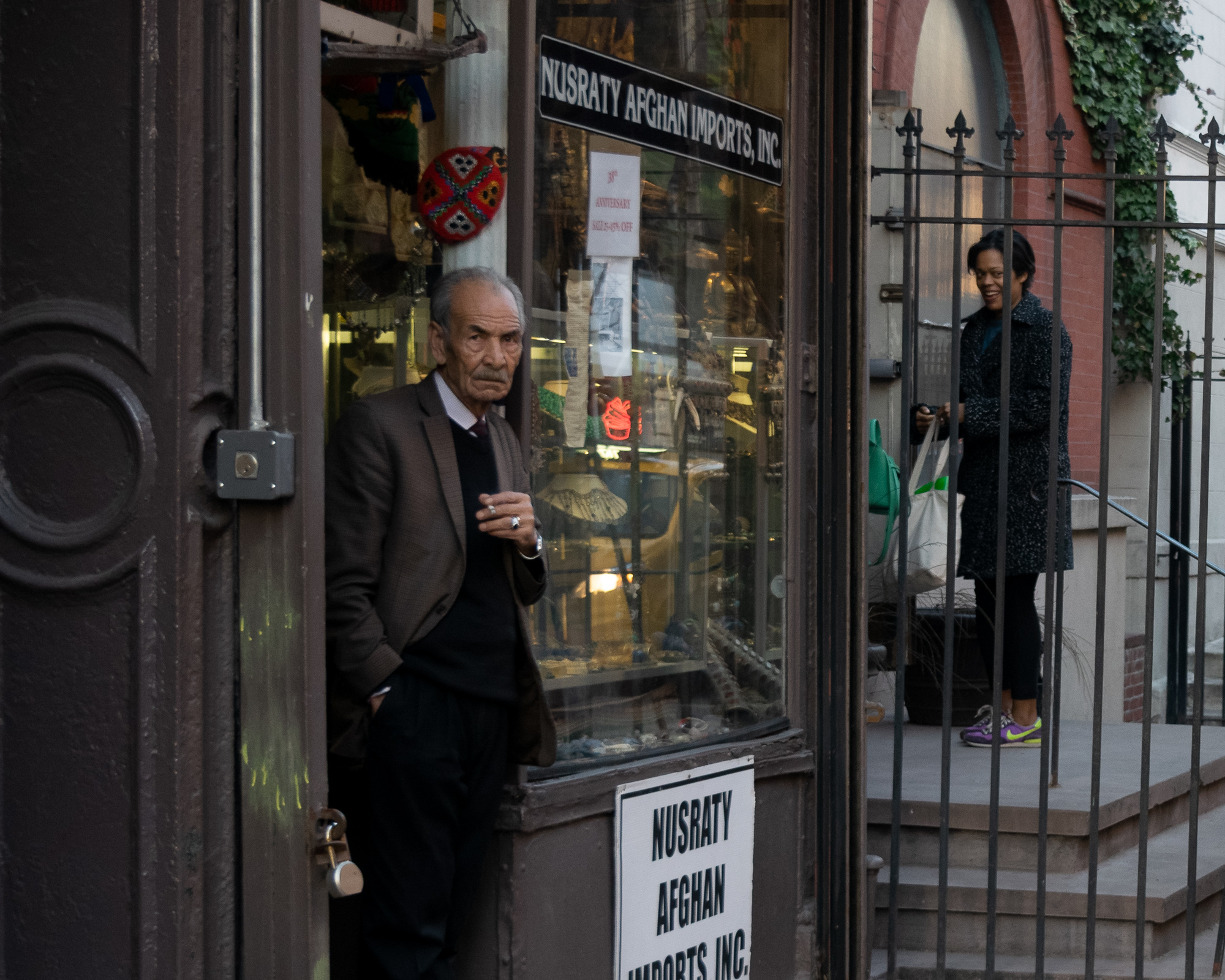 An older man in a brown jacket and black sweater stands in the open doorway of Nusraty Afghan Imports, holding a cigarette and looking directly at the camera, while a woman in a dark tweed coat carrying tote bags stands behind an iron fence on the stoop next door.