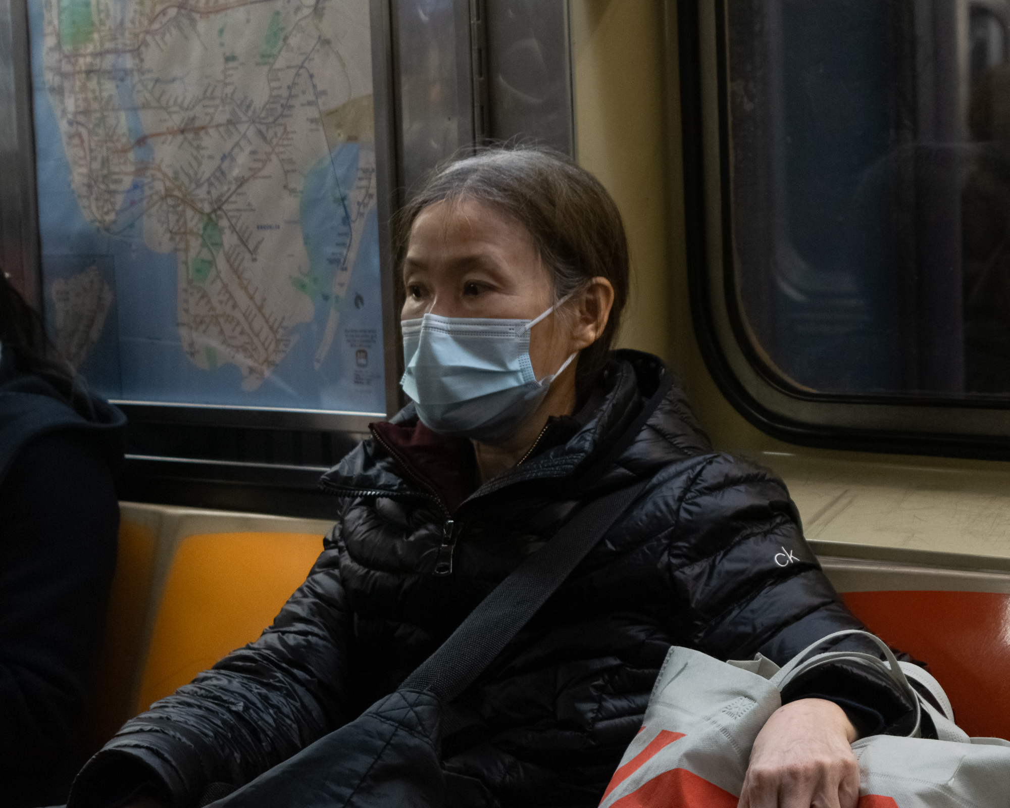 A woman in a black quilted Calvin Klein puffer jacket and a light blue surgical mask sits on a subway bench, looking off to her left, with a laminated NYC transit map visible in the window behind her. A canvas tote bag rests across her lap and her hand rests on her knee, with an orange and red seat visible beside her.
