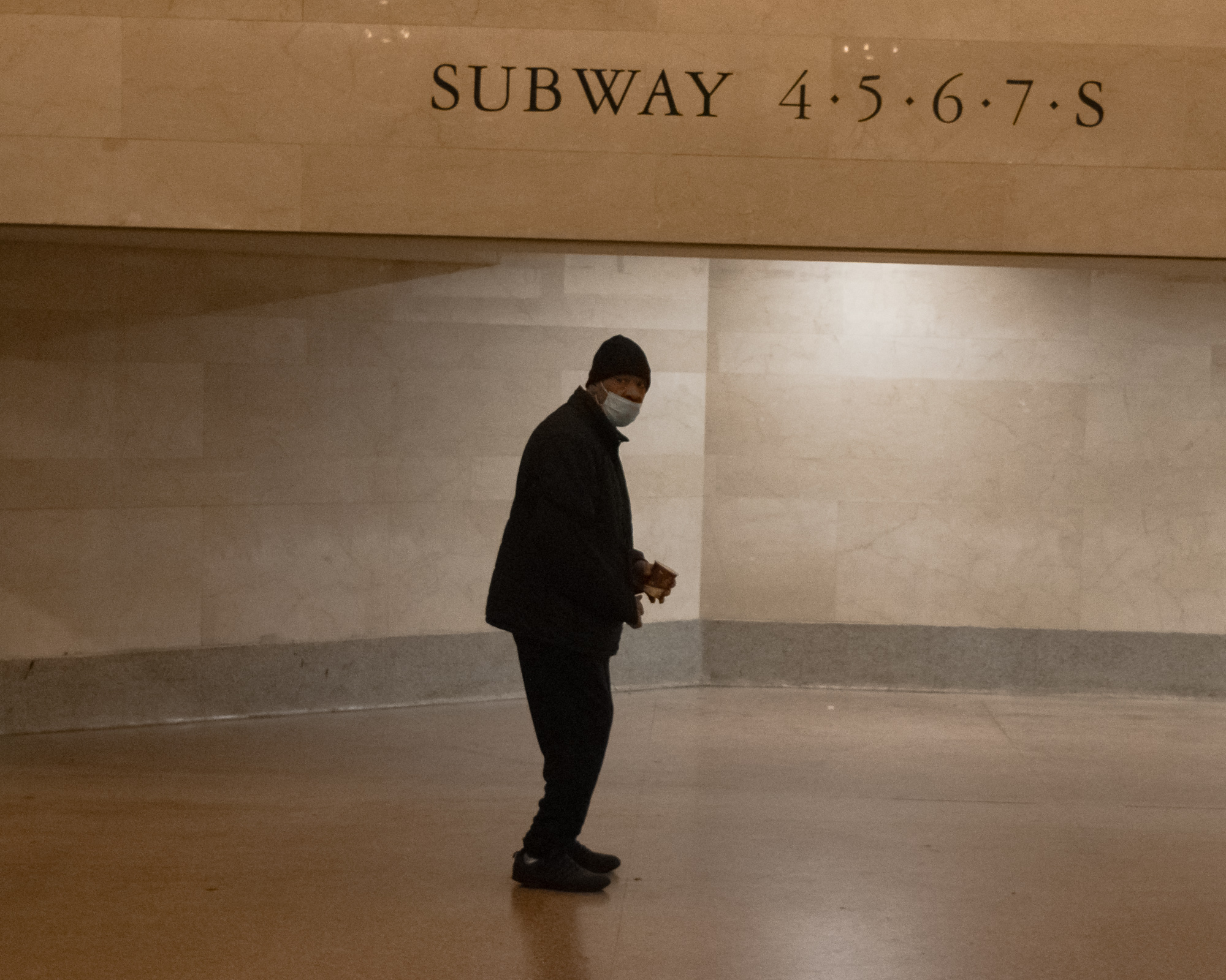 A man in a dark winter coat, black knit cap and surgical mask stands alone on a polished floor, turning to look toward the camera while holding a small brown paper bag. Above him, carved limestone lettering reads SUBWAY 4 5 6 7 S.