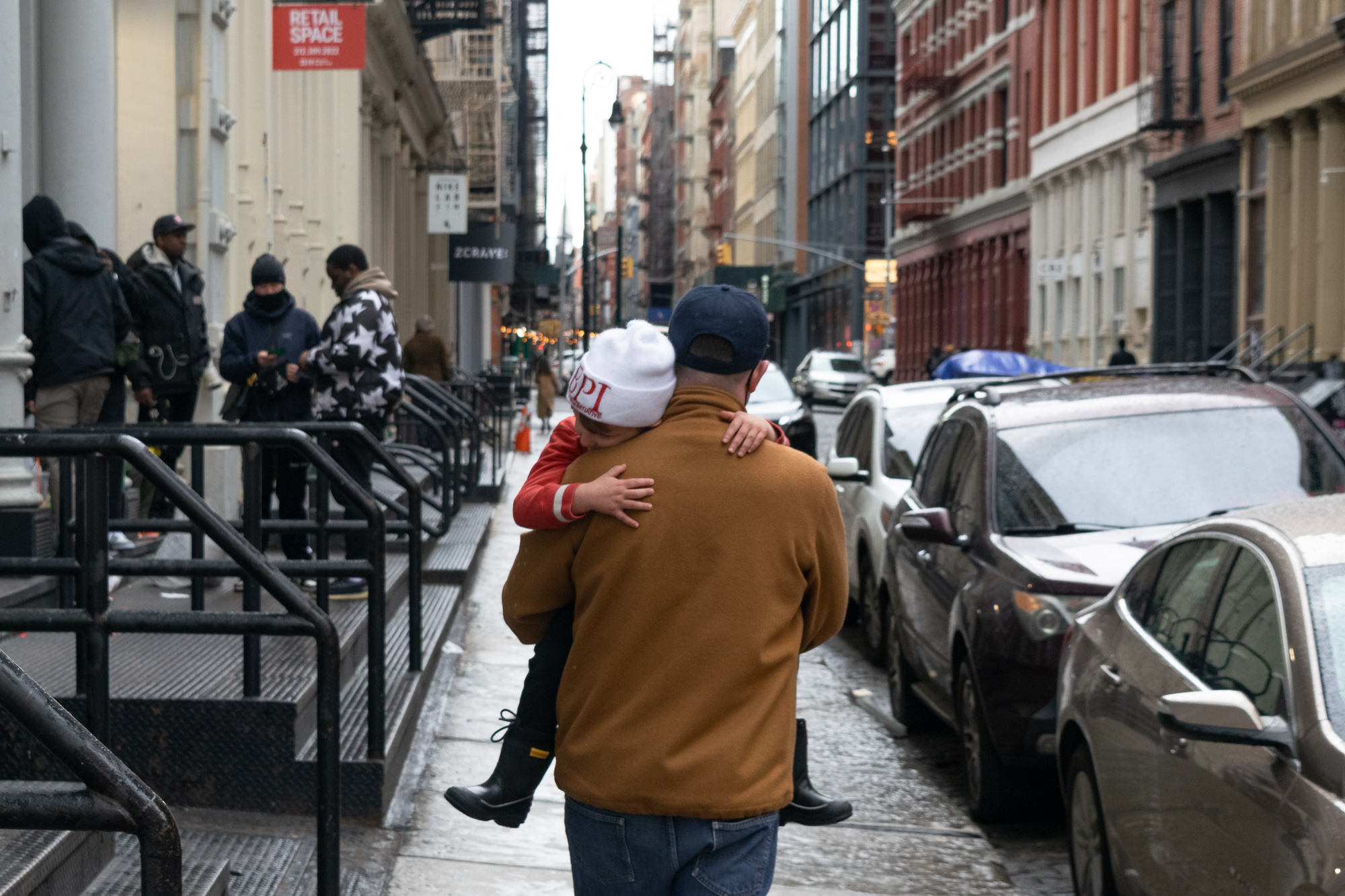 A man in a tan fleece jacket and navy baseball cap carries a child on his hip along a wet Manhattan sidewalk; the child, in a white knit beanie and red jacket with black rain boots, rests their head on his shoulder with an arm around his neck. Cast-iron loft buildings, parked cars and a group of people standing near a stoop fill the background.