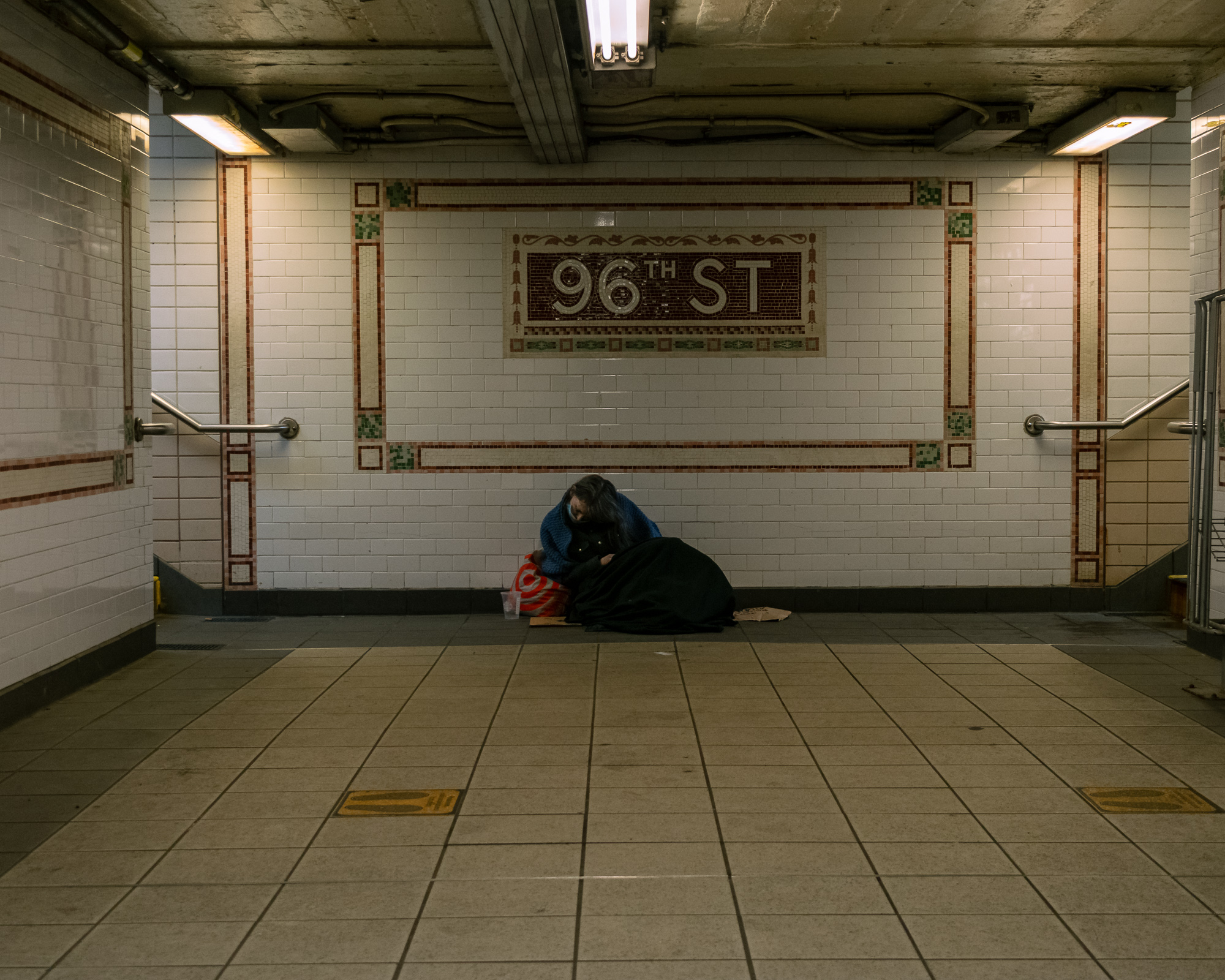 In a nearly empty subway mezzanine, a woman in a blue knit sweater and face mask sits on the tiled floor against a white tiled wall, bent over a red Target bag and a black blanket. Above her, a large ceramic mosaic reads 96TH ST, flanked by stair handrails and fluorescent ceiling lights.