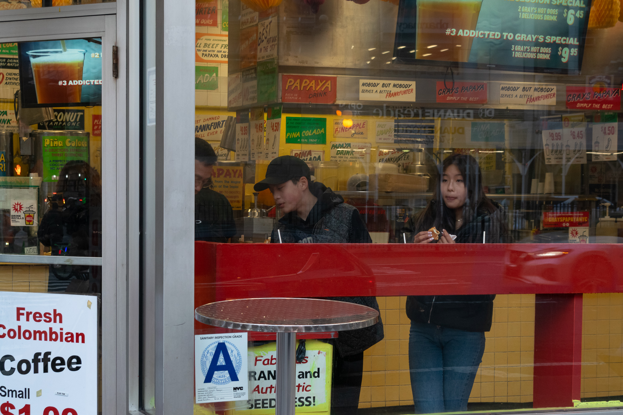 Seen through a storefront window, two teenagers stand at a red window counter inside Gray's Papaya. A boy in a black cap and gray hooded jacket looks down, while a girl in a dark jacket and jeans holds a partly eaten hot dog. Hand-lettered signs for papaya drink, pina colada and frankfurter specials cover the yellow tiled wall behind them.