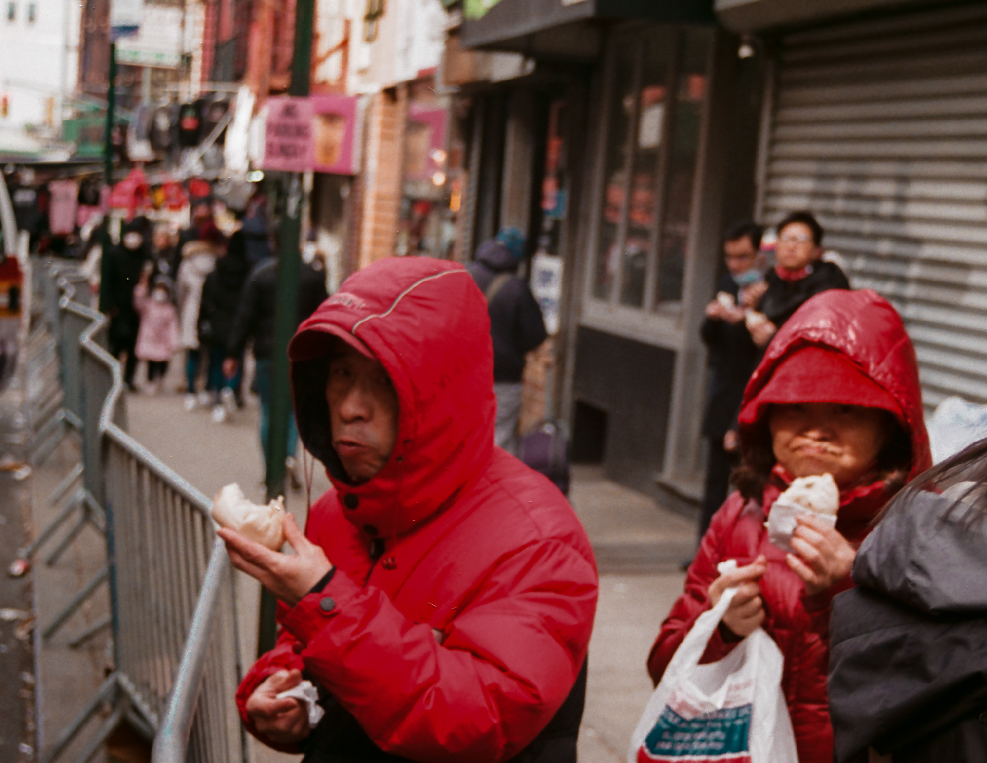 A man in a bright red hooded winter coat stands on a Chinatown sidewalk mid-bite into a steamed bun, with a woman in a matching red hood eating beside him and holding a white plastic grocery bag. Behind them, metal barricades line the curb and pedestrians pass a shuttered storefront.