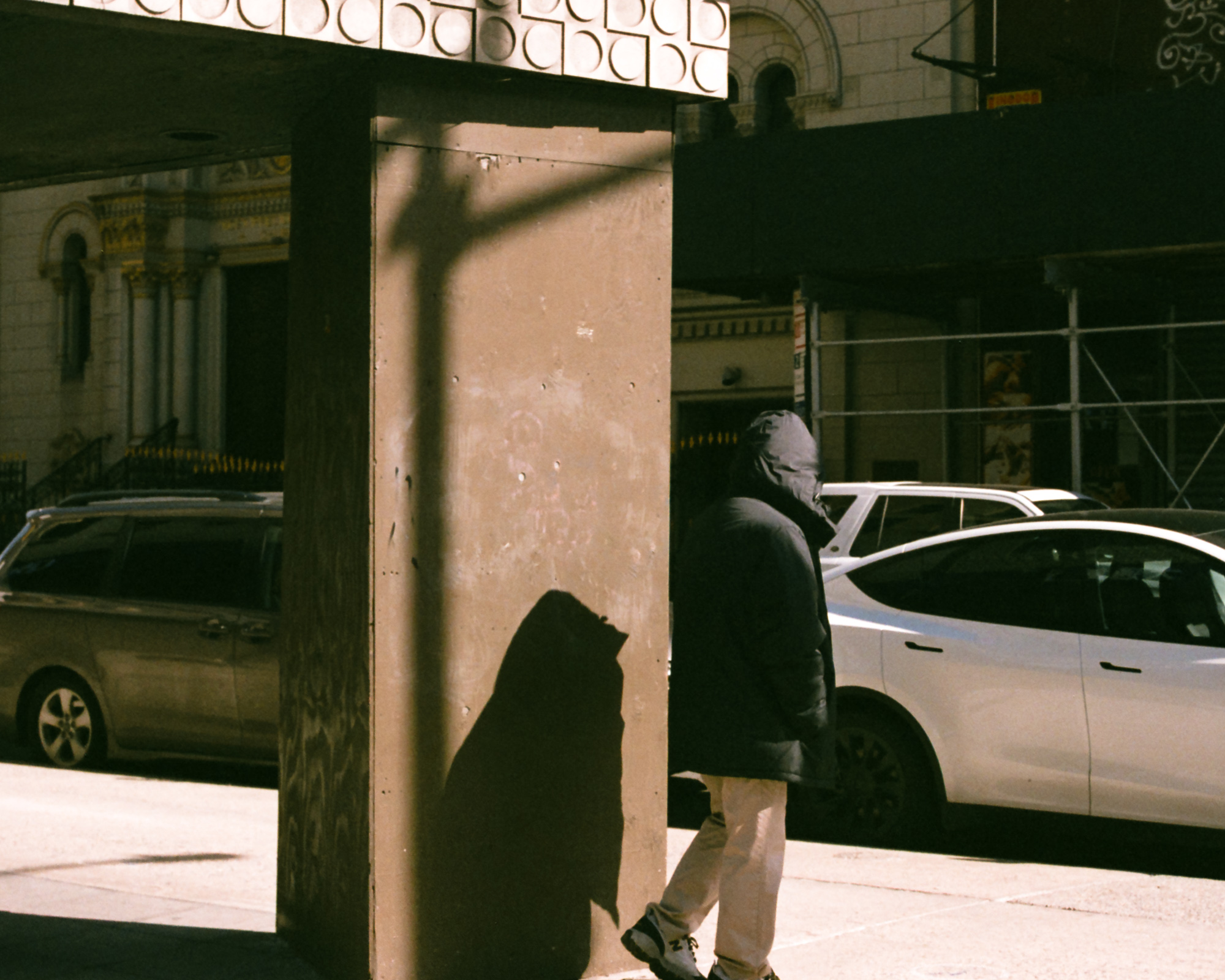 A person in a hooded black puffer coat and light pants walks past a plywood-clad scaffolding column on a sunlit Manhattan sidewalk, their dark silhouette thrown across the boards. Behind them a silver sedan and a minivan sit parked in front of an ornate stone building.